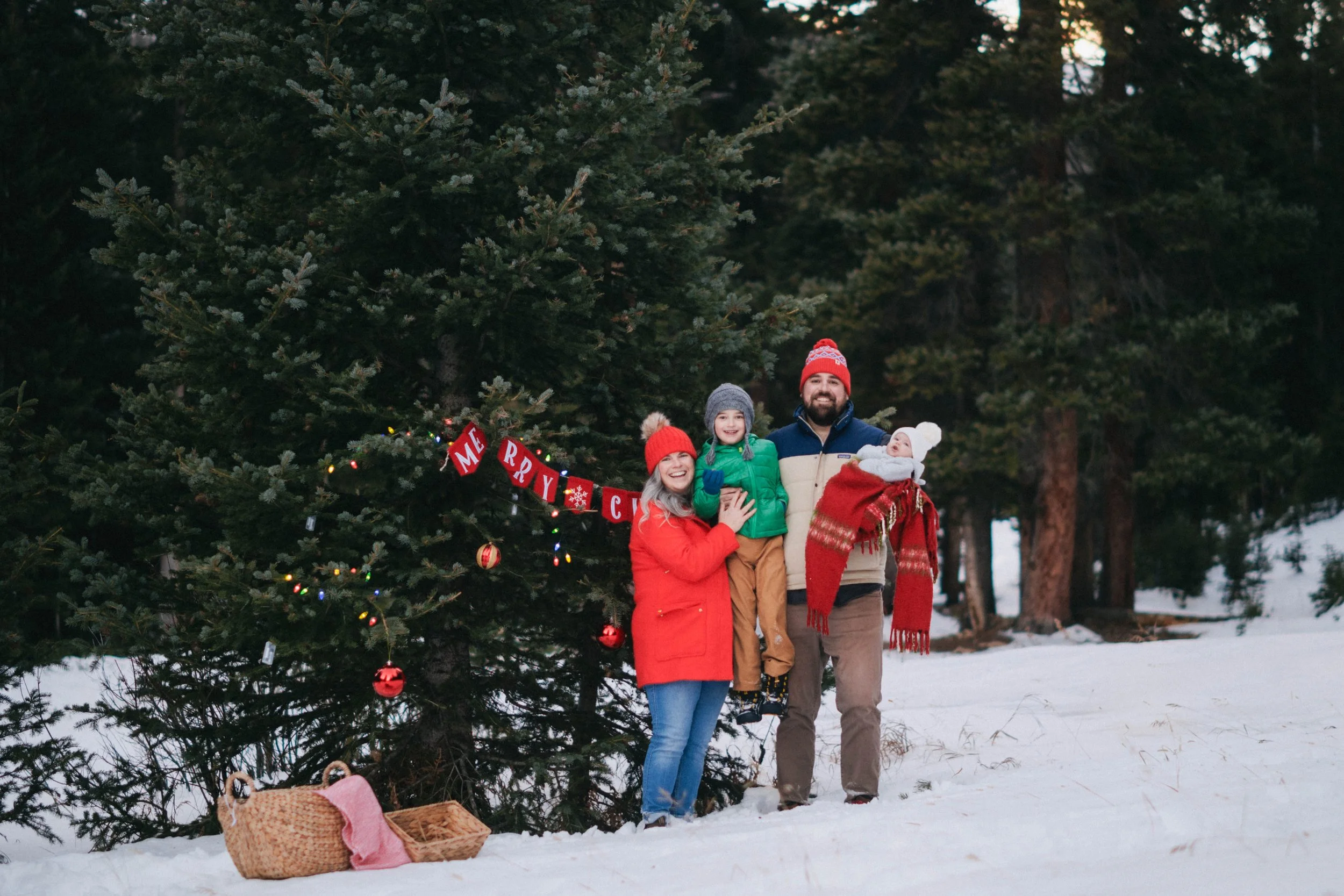 Family posing in front of a decorated Christmas tree outdoors, with snow on the ground and a snowy forest background.