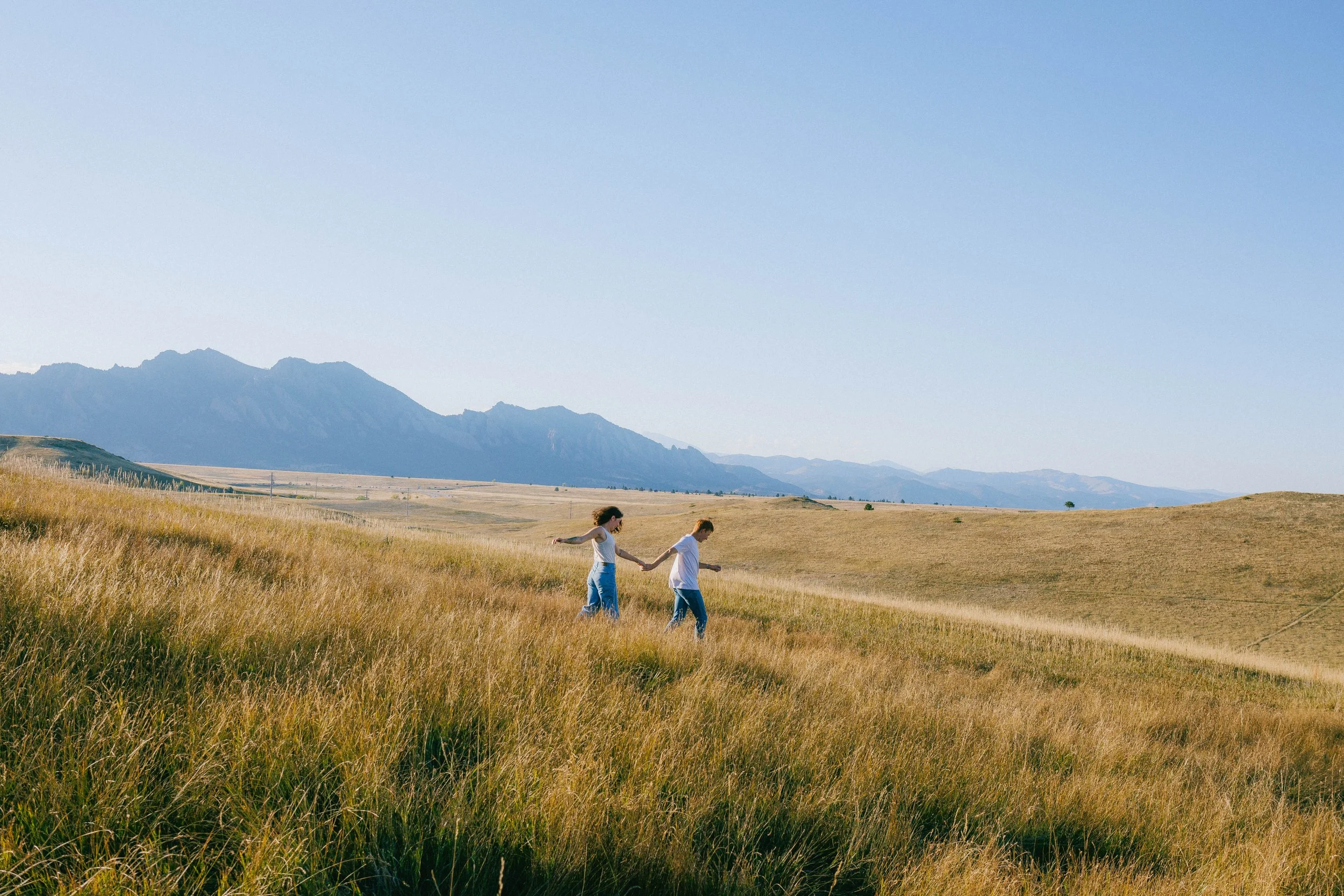 Engagement and couples portrait walking hand in hand in a grassy field with mountains in the background.