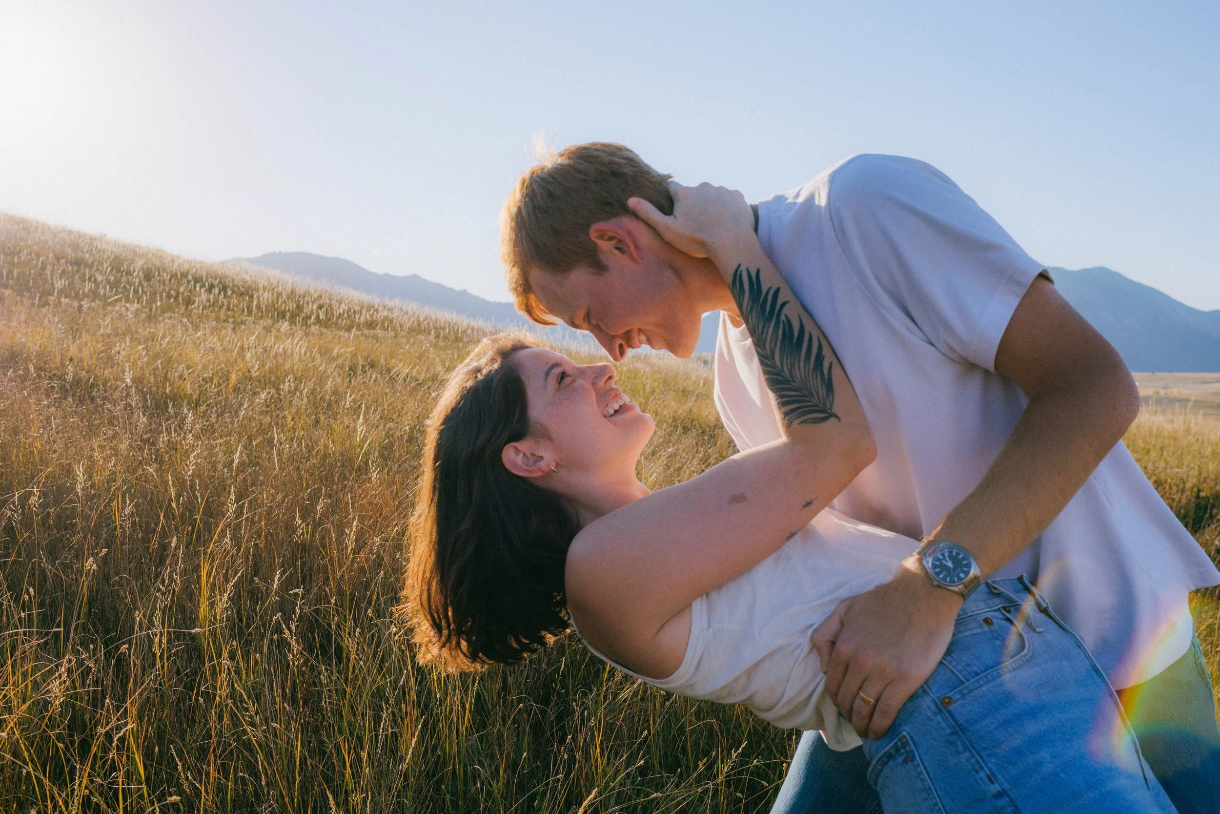 Couples portrait photo embracing in a grassy field with mountains in the background.