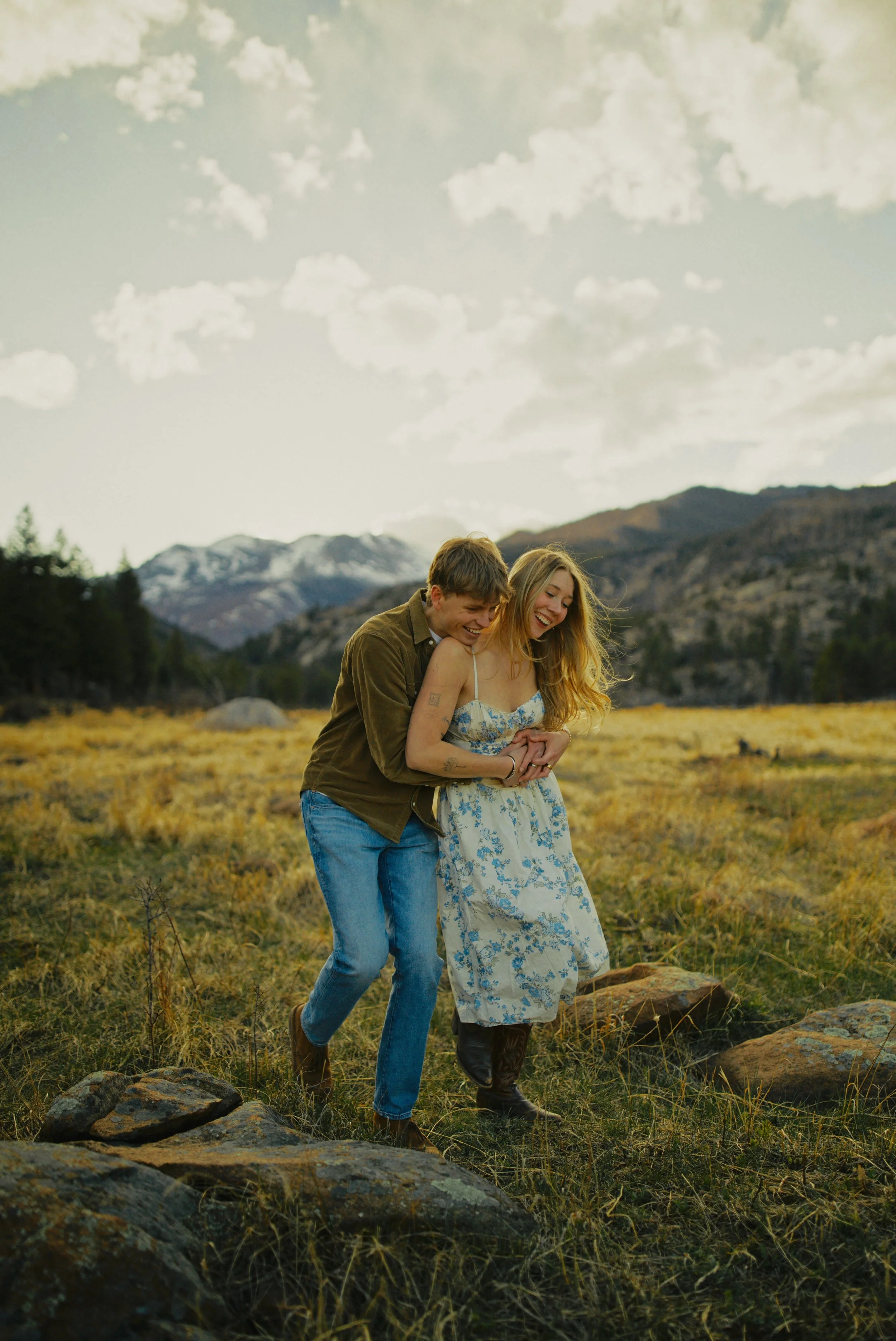A smiling young couple laugh and walk hand-in-hand through a mountainous field with rocks and tall grass, with a mountain range and cloudy sky in the background, during late afternoon or early evening.