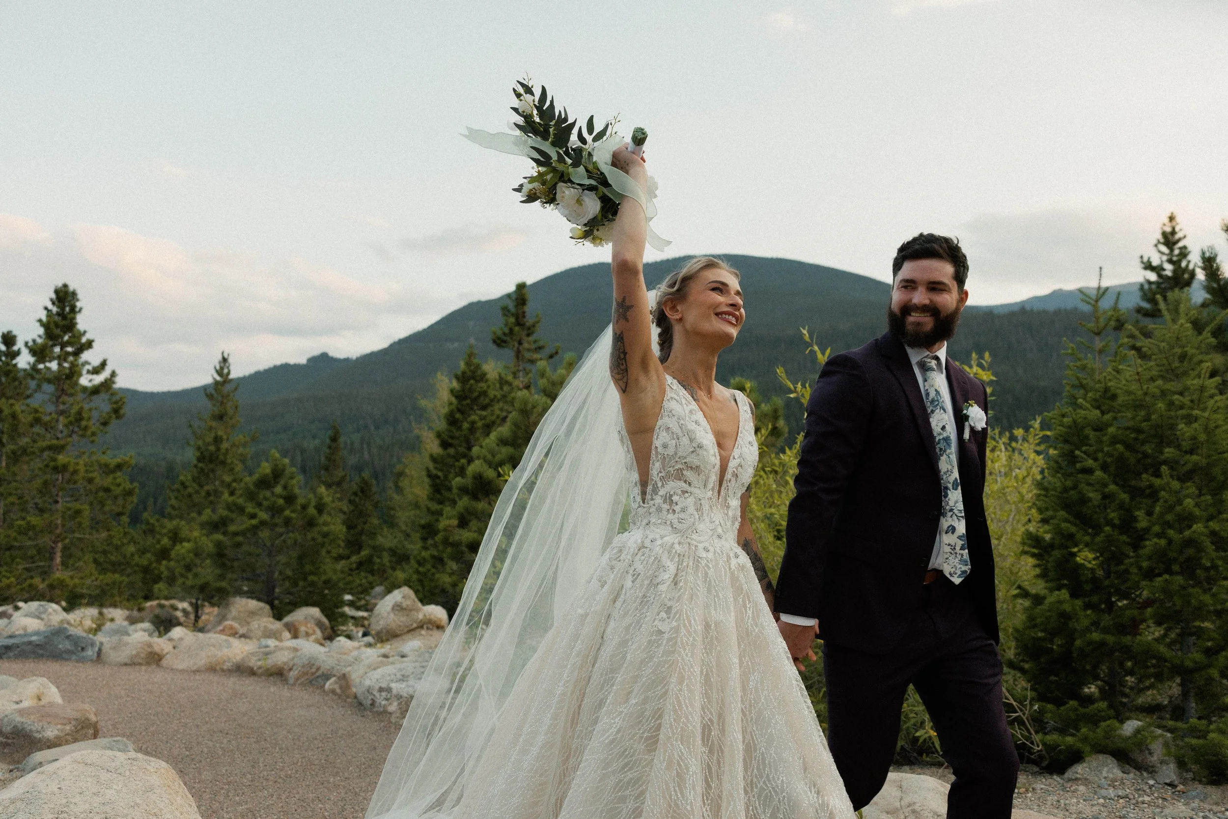 A bride and groom walk outdoors in a forested mountainous area, holding hands. The bride is smiling, wearing a lace wedding dress and holding a bouquet, while the groom is dressed in a dark suit and floral tie, with a smile.