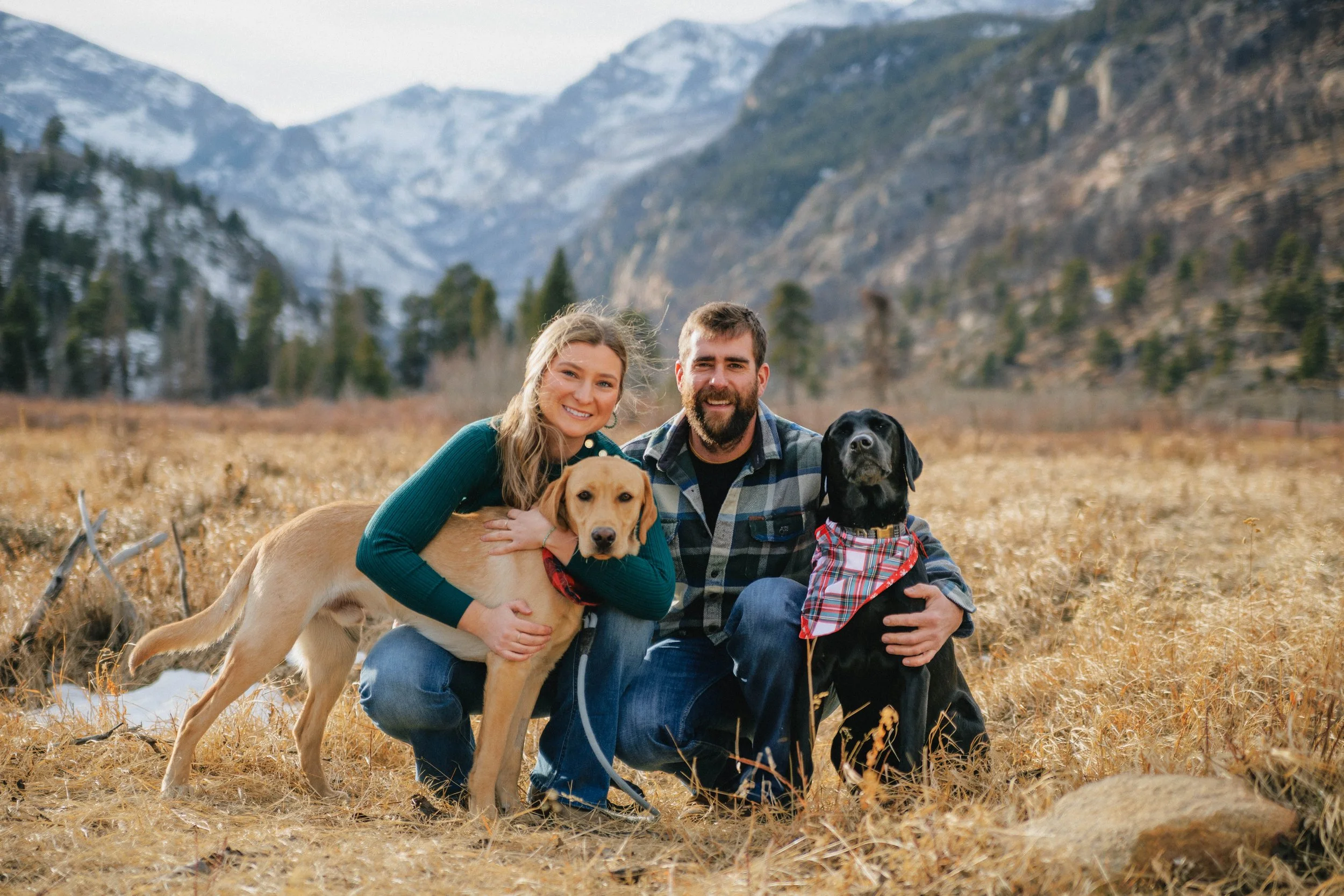 A couple kneeling in a field with two dogs, one tan and the other black wearing a red plaid scarf, against a mountainous backdrop.