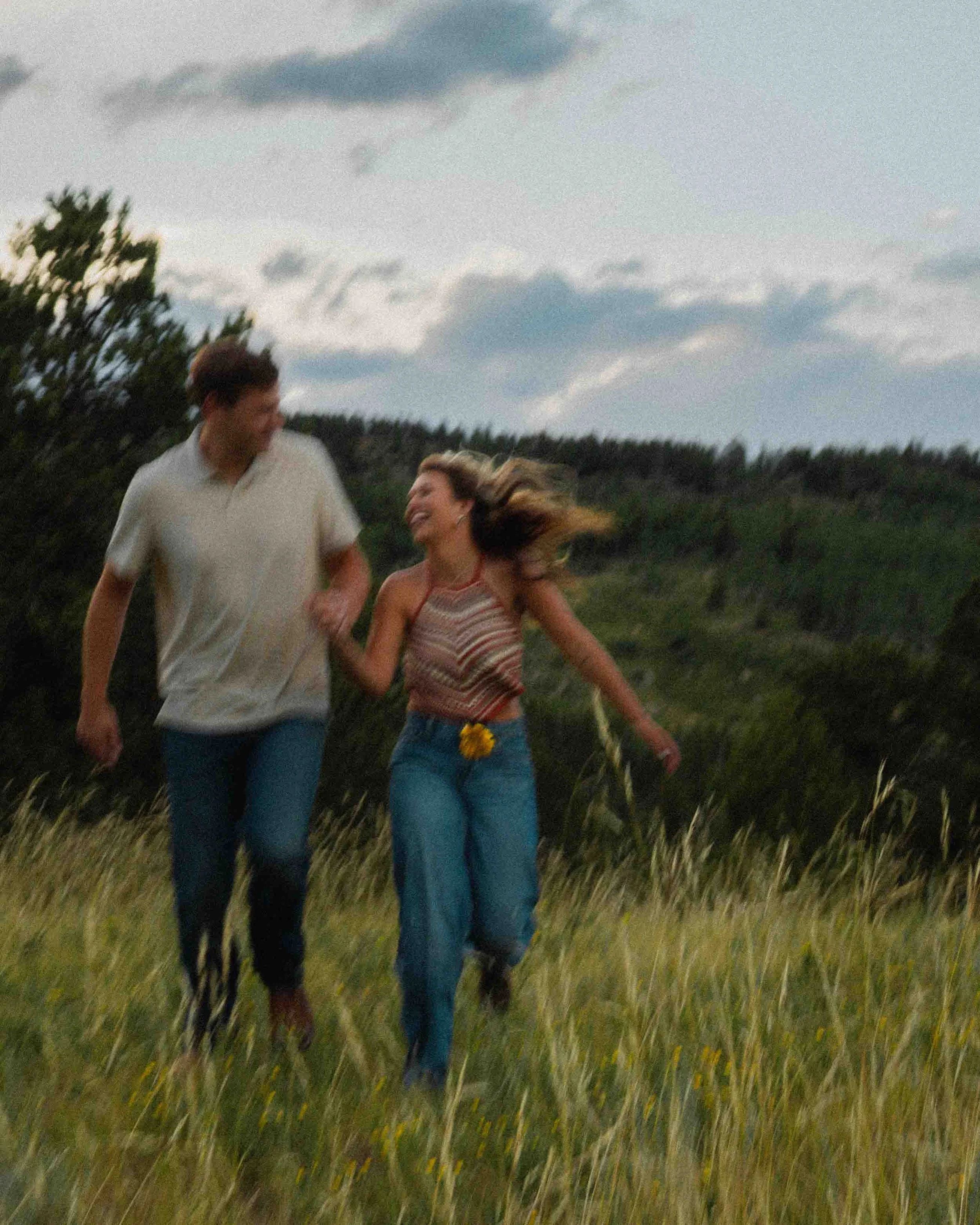 A young couple running hand in hand through a grassy field during daytime, smiling and enjoying each other's company.