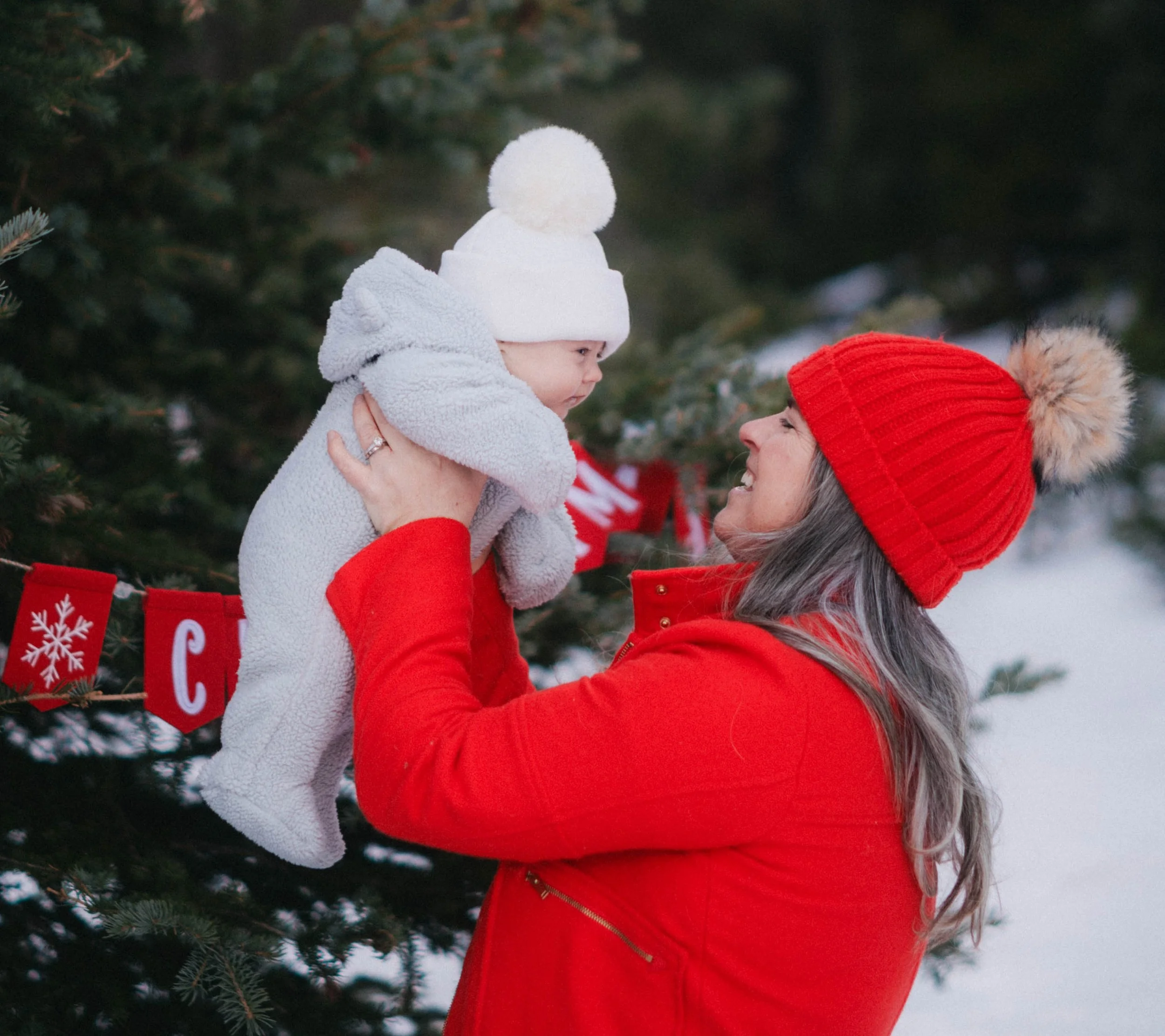 Woman in red coat and hat holding a baby in gray outfit and white hat outdoors, with a festive Christmas banner and trees in the background.