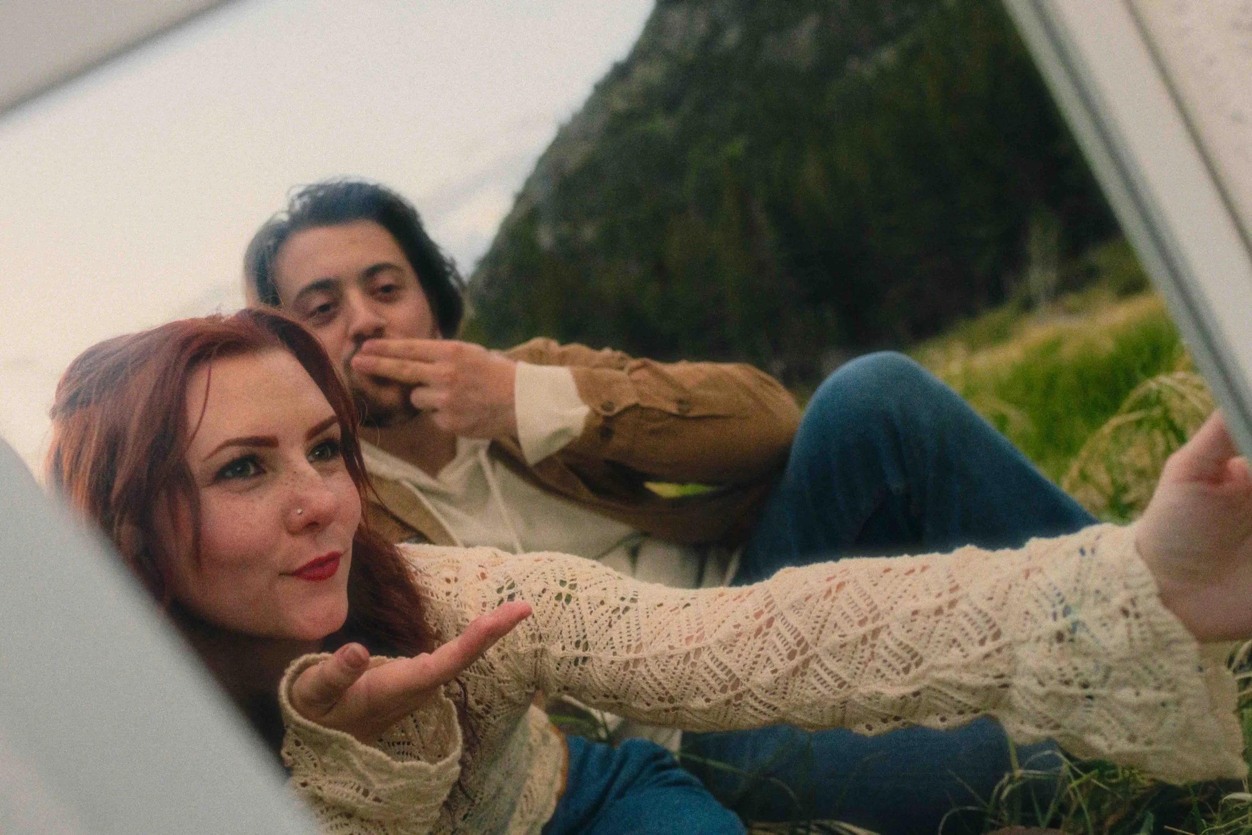 Two people lying on grass outdoors, taking a selfie. The woman in front has red hair, freckles, and a nose piercing, wearing a cream-colored crochet sweater. The man behind her has dark hair, wearing a brown jacket and white shirt. They are surrounde