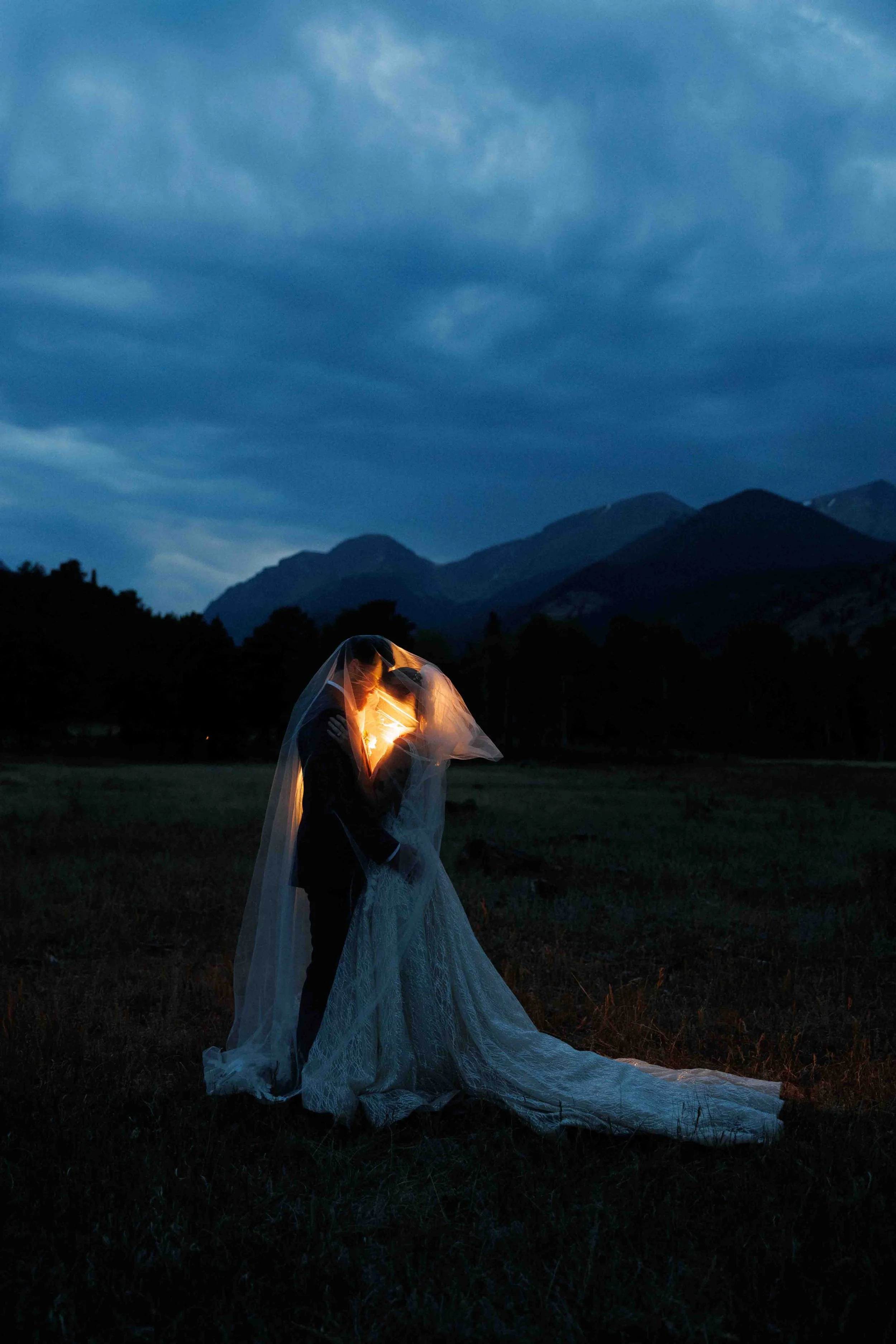 A couple dressed in wedding attire, sharing a kiss under a transparent veil at dusk or night, with a mountainous landscape and dark sky in the background.