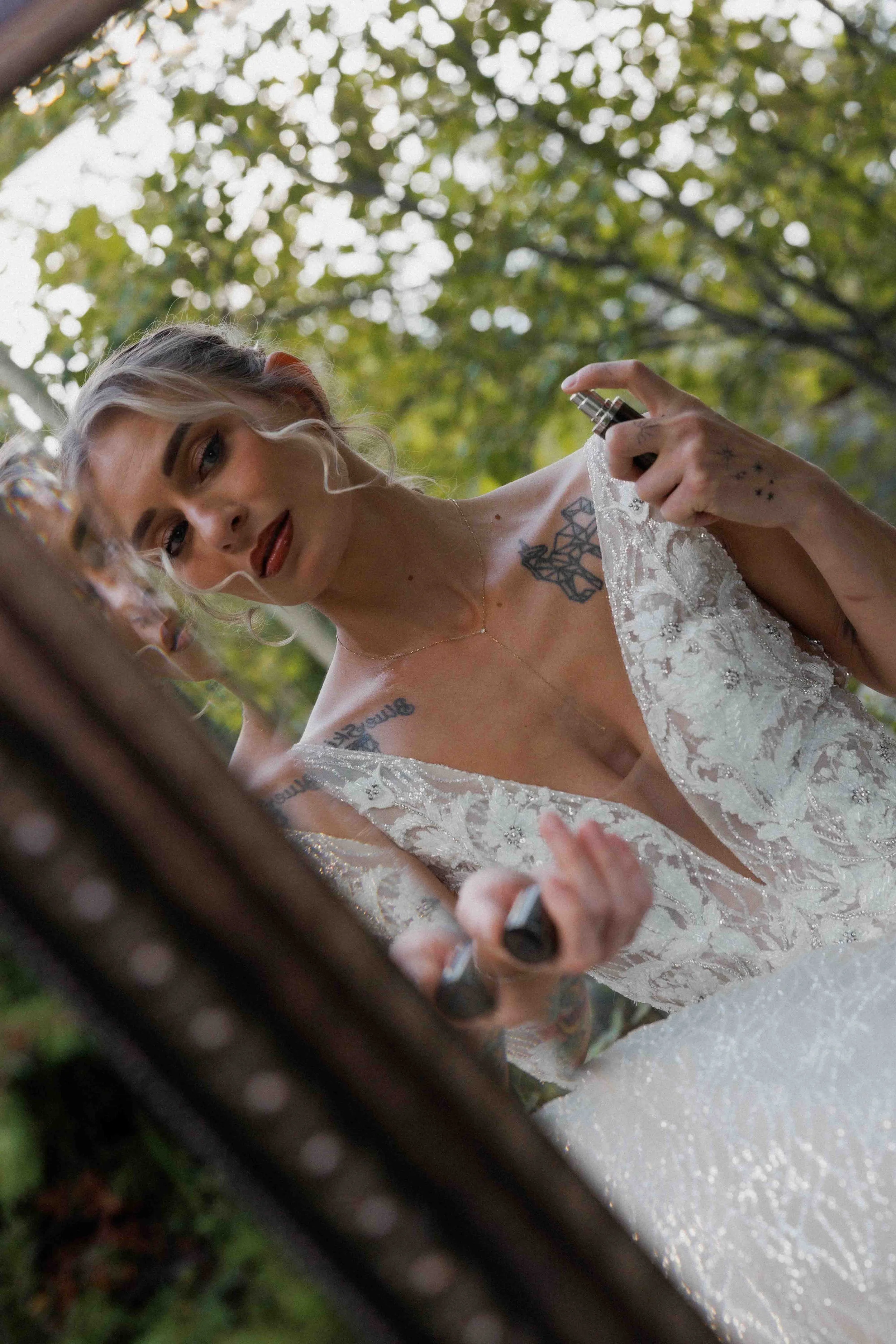 A woman with blonde hair, tattoos, and makeup in a wedding dress applies lipstick while looking in a mirror outdoors with green trees in the background.
