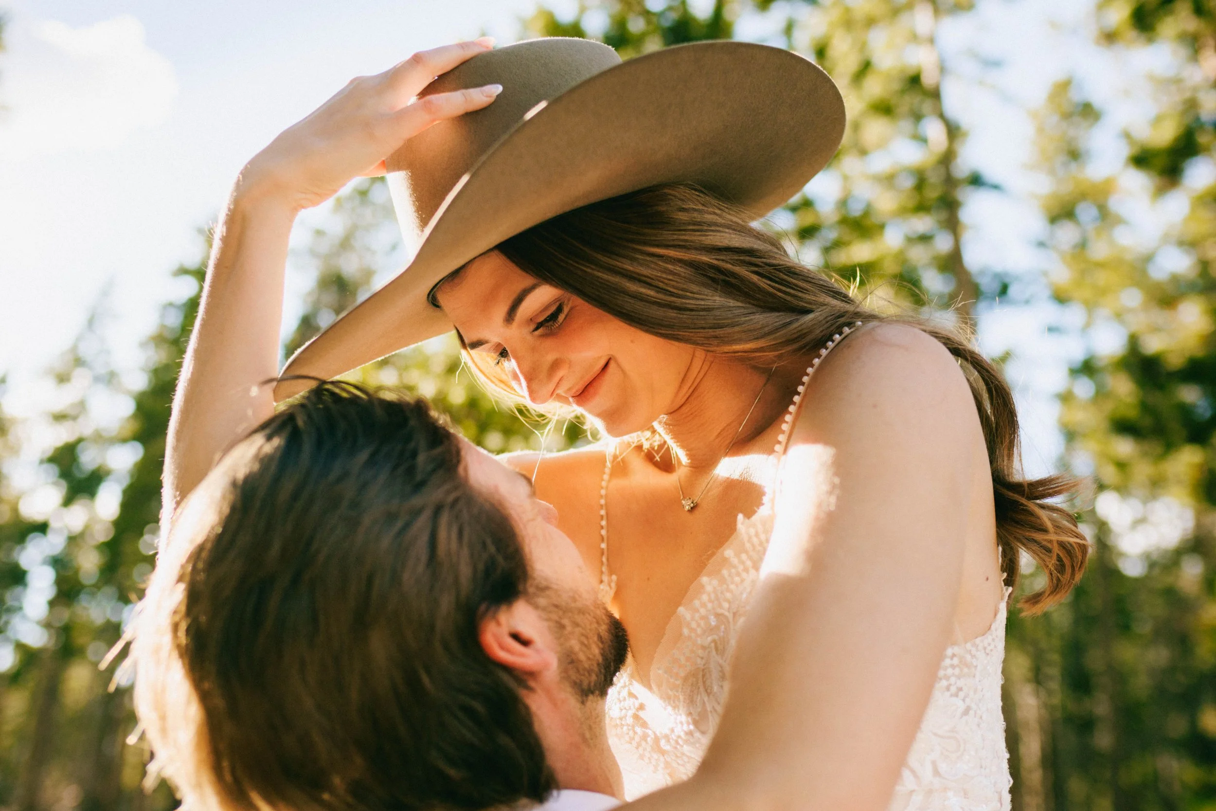 Couple embracing outdoors, woman in hat smiles at man against tree background. Engagement photo wedding elopement