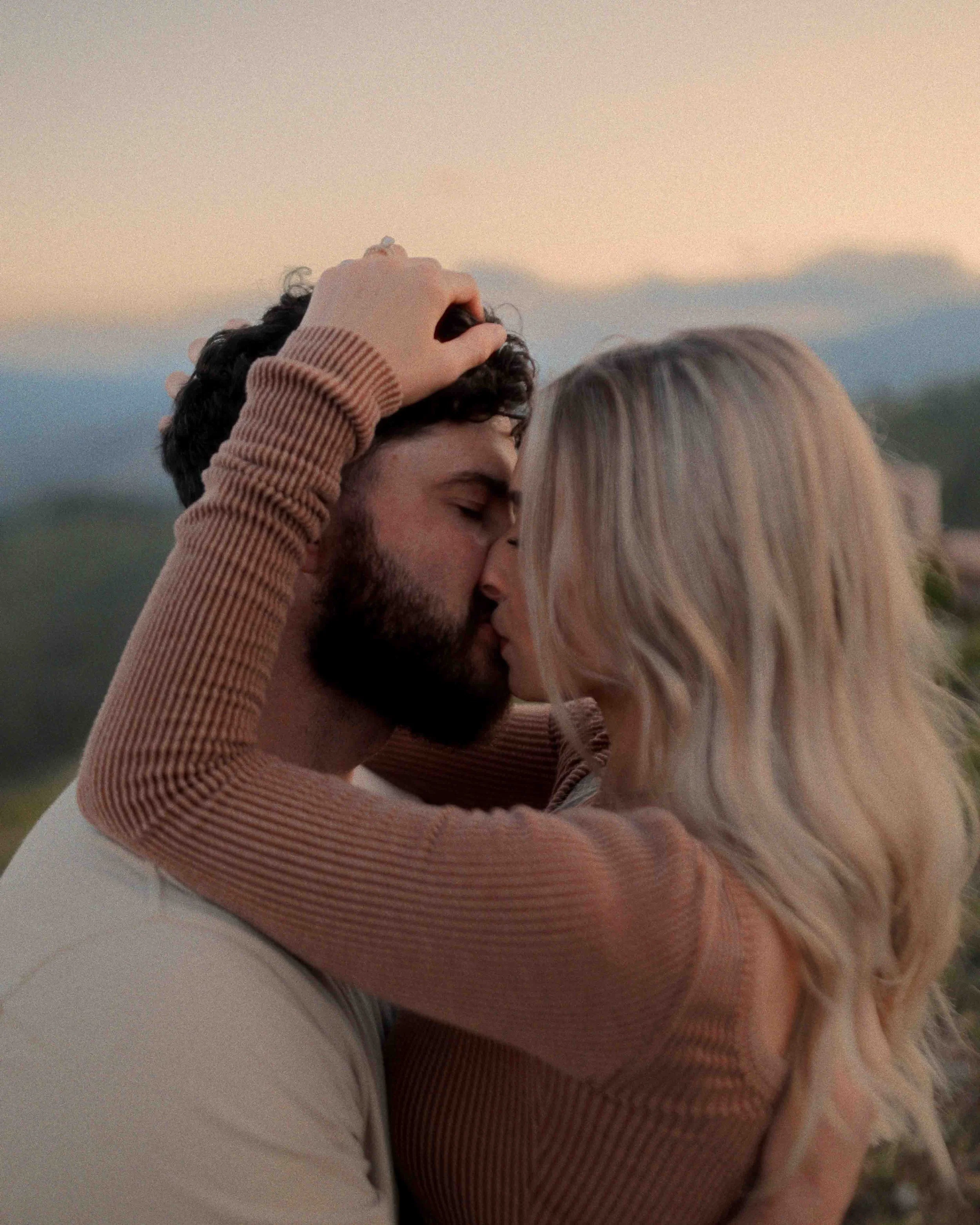 A couple sharing a kiss outdoors at sunset, with the man's hand in the woman's blonde hair.