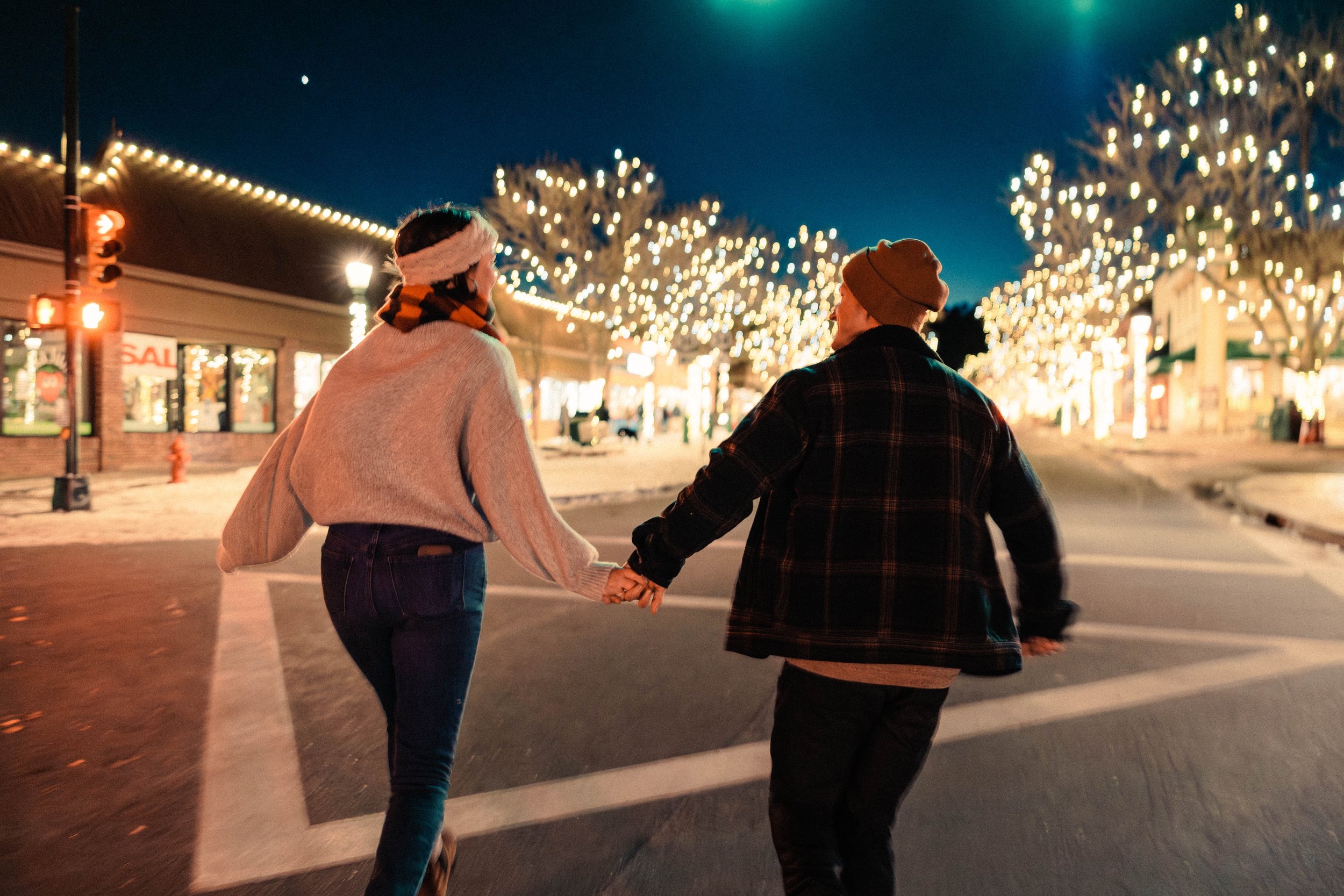 A couple holding hands and walking at night on a street with festive lights. The street is decorated with string lights on trees and buildings. Both are wearing winter clothing, including a beanie and scarf.