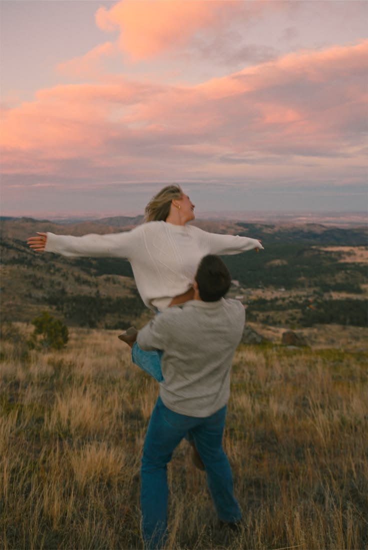 A man lifting a woman with outstretched arms in an open field during sunset with pink and orange clouds in the sky.