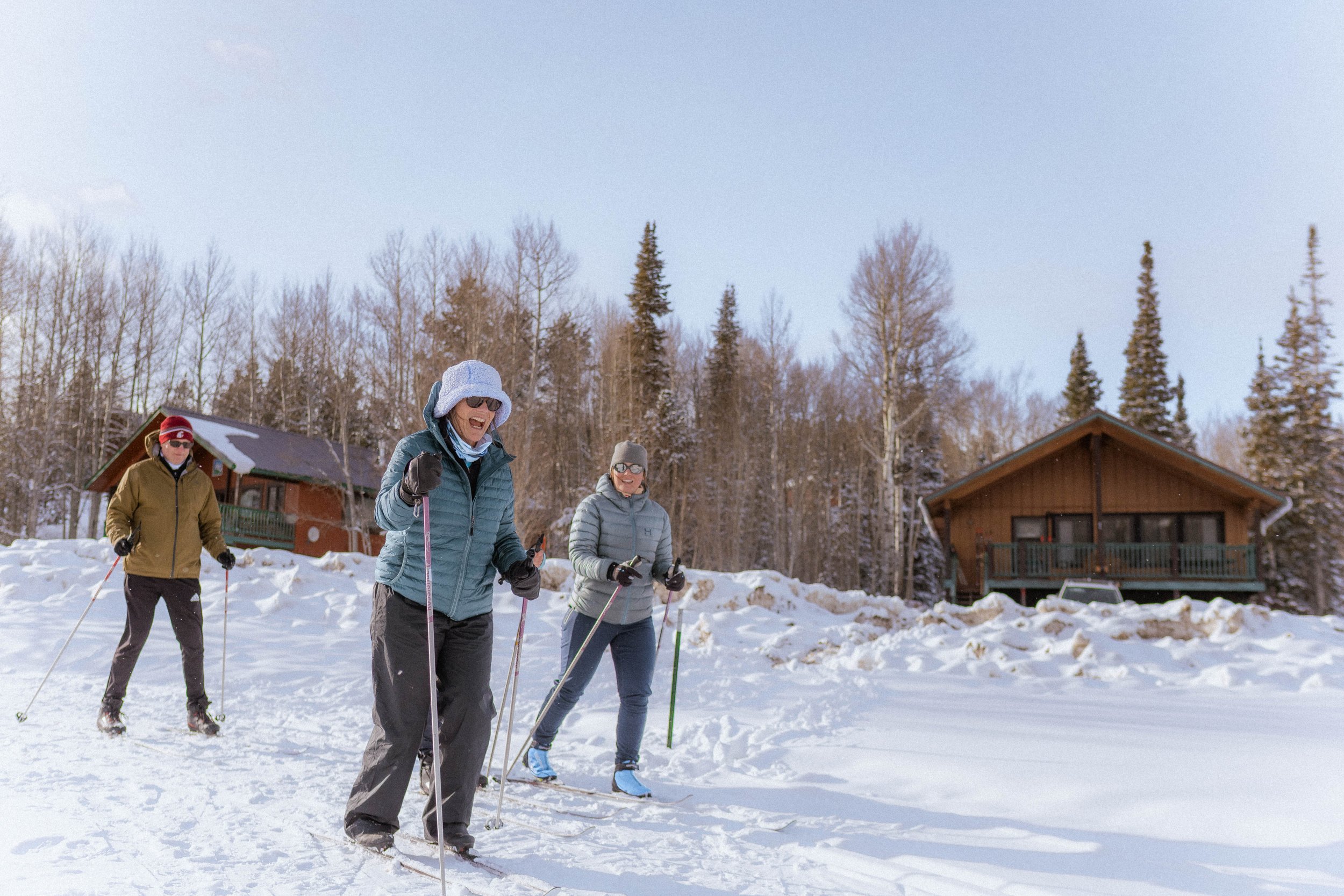 Three people cross-country skiing in snowy landscape with cabins and trees in the background.