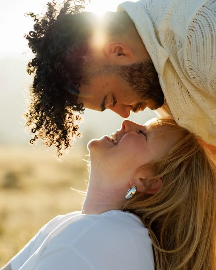 A couple sharing a close moment outdoors, faces touching, with sunlight shining behind them.