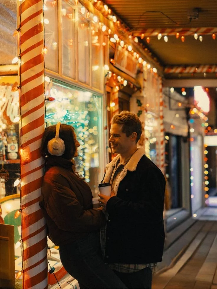 A man and woman standing outside a decorated street shop at night, surrounded by Christmas lights in Estes. The woman is wearing earmuffs and a warm coat, leaning against a candy cane-striped pole, while the man holds a coffee cup and smiles at her.