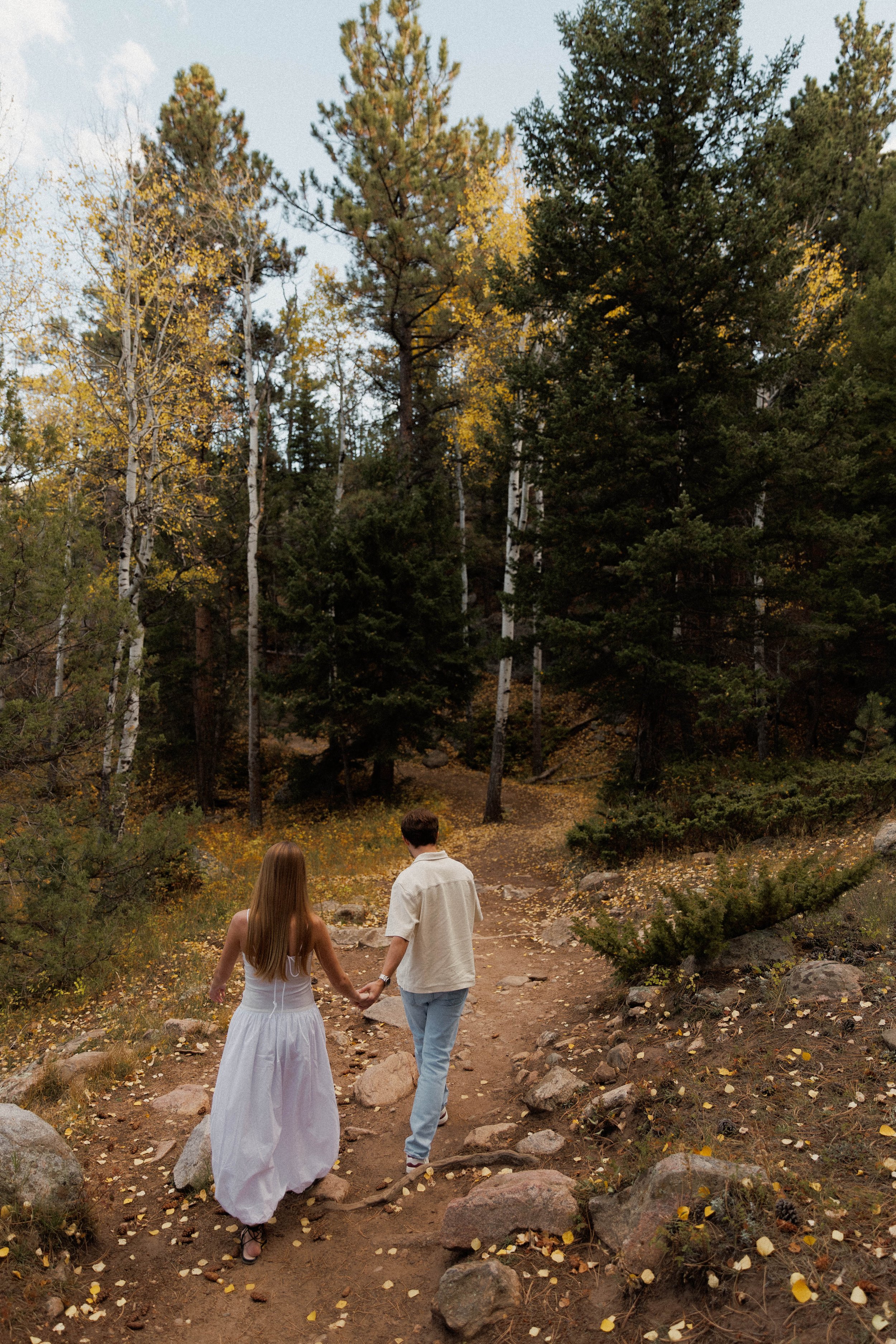 A couple holding hands and walking on a dirt trail surrounded by trees in an autumn forest.