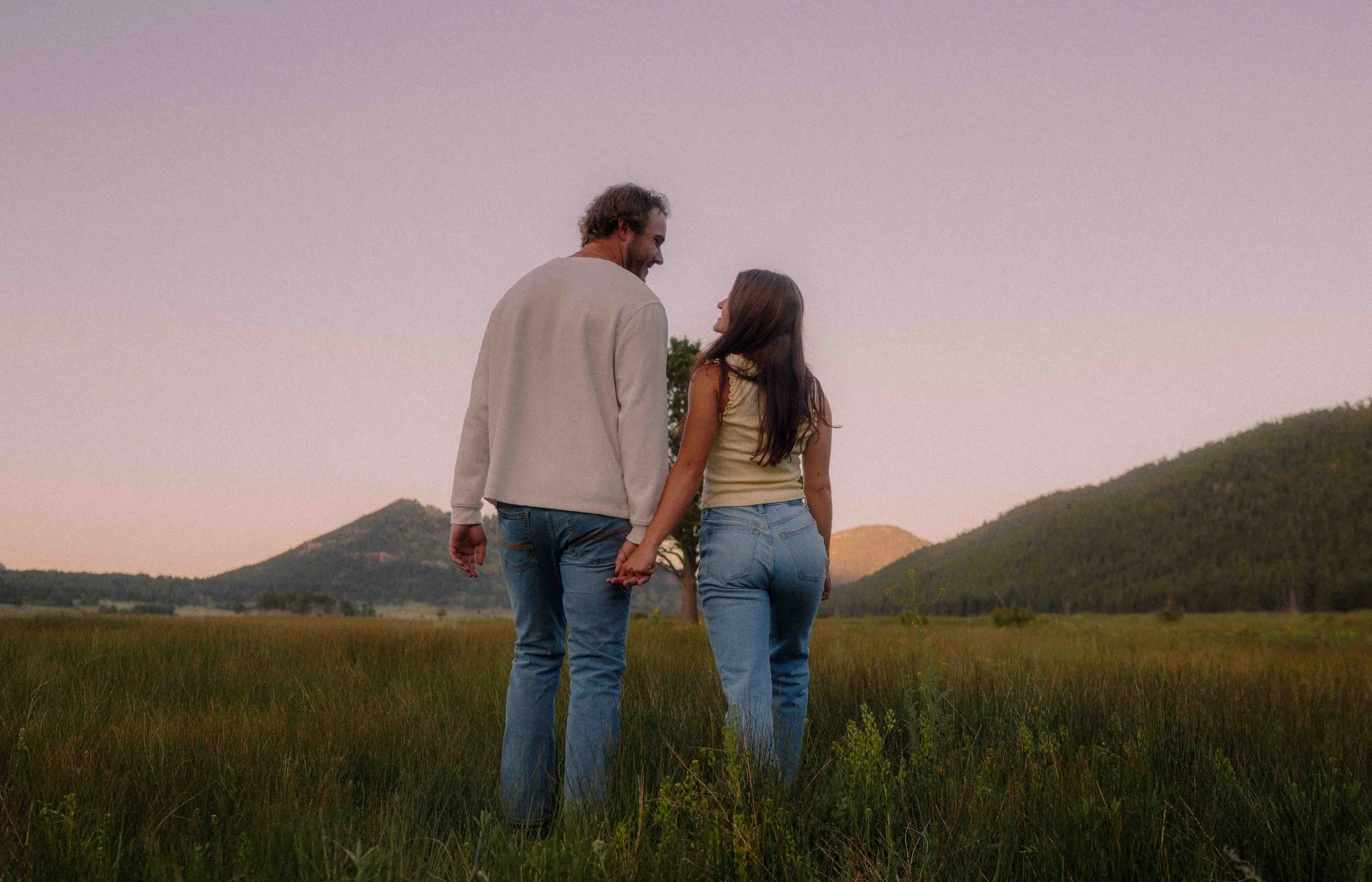 A man and woman holding hands and walking in a grassy field with hills in the background during sunset.