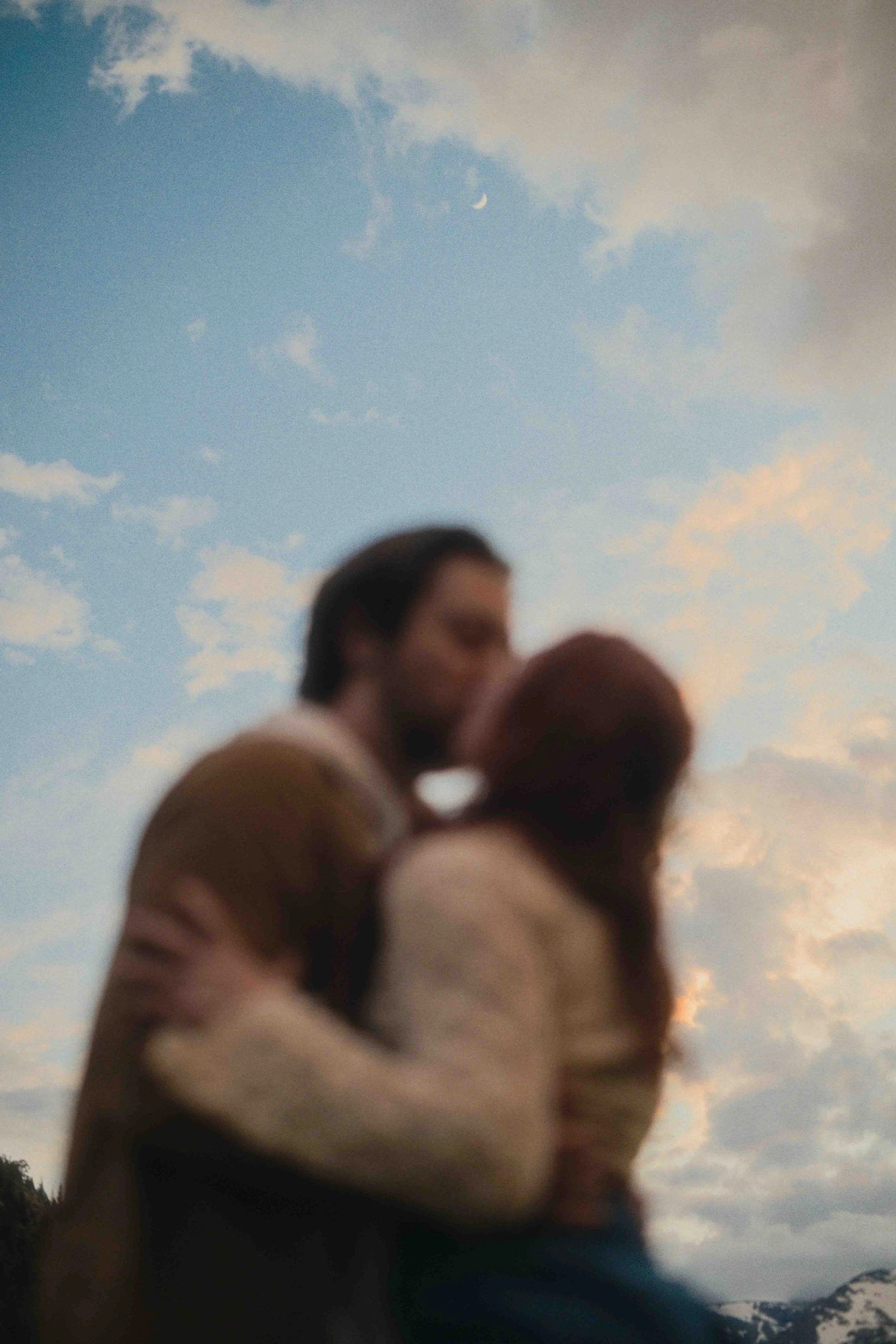 Blurred image of a couple kissing outdoors under a sky with clouds and a visible crescent moon.