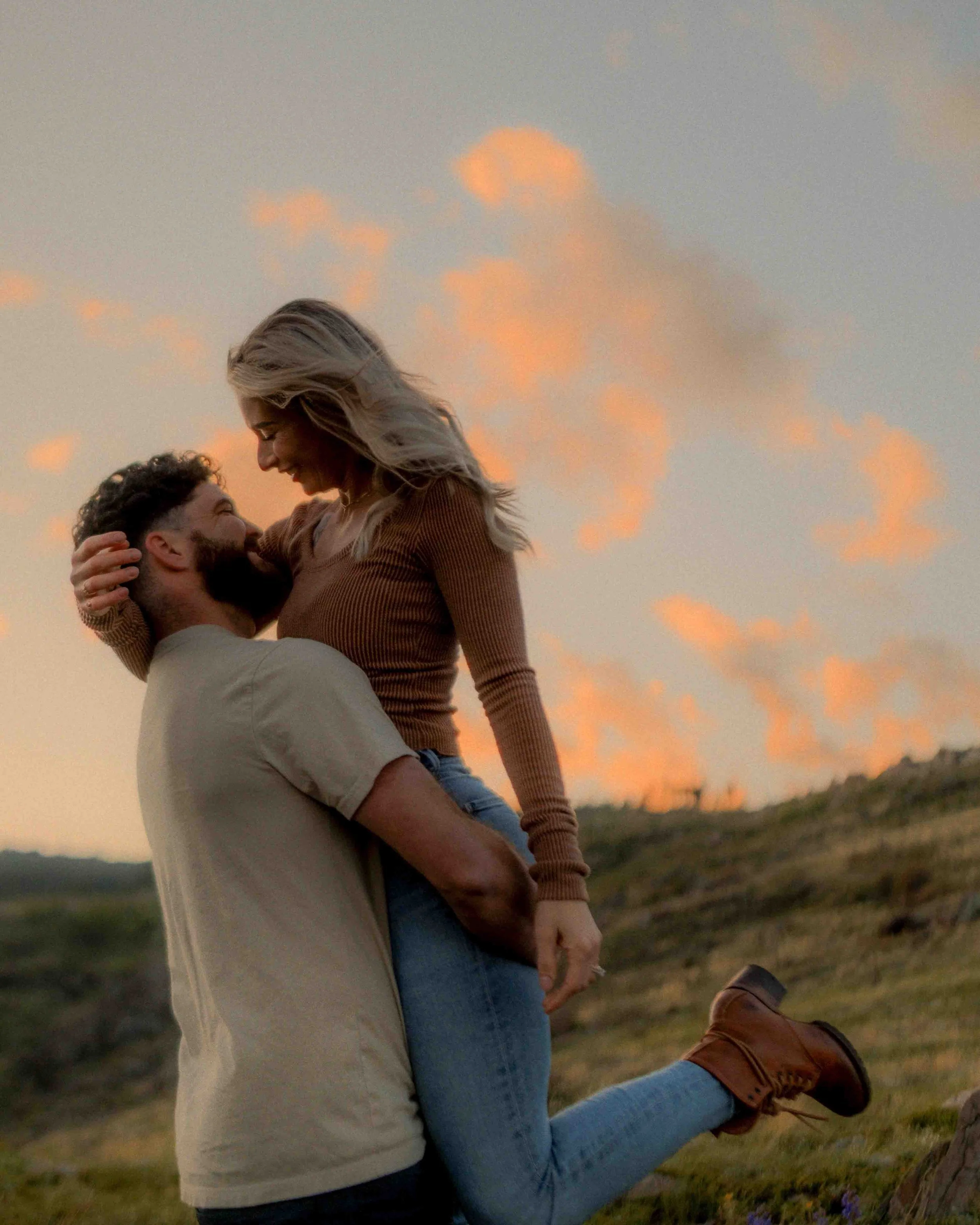 A couple outdoors at sunset, with the man lifting the woman, both smiling and looking at each other, in a natural landscape.