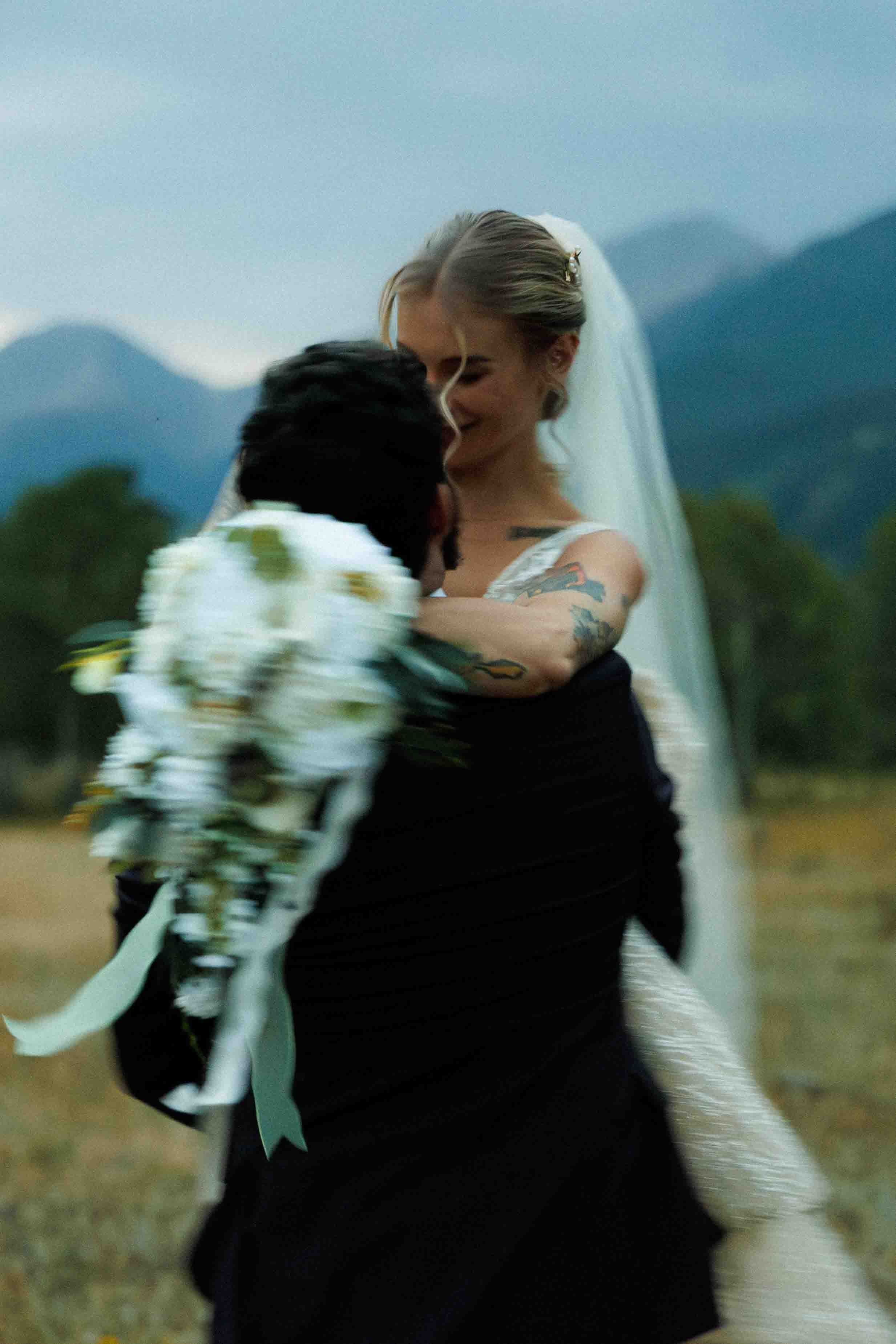 A bride and groom embracing outdoors during their wedding, with mountains in the background and the bride holding a bouquet of white flowers.