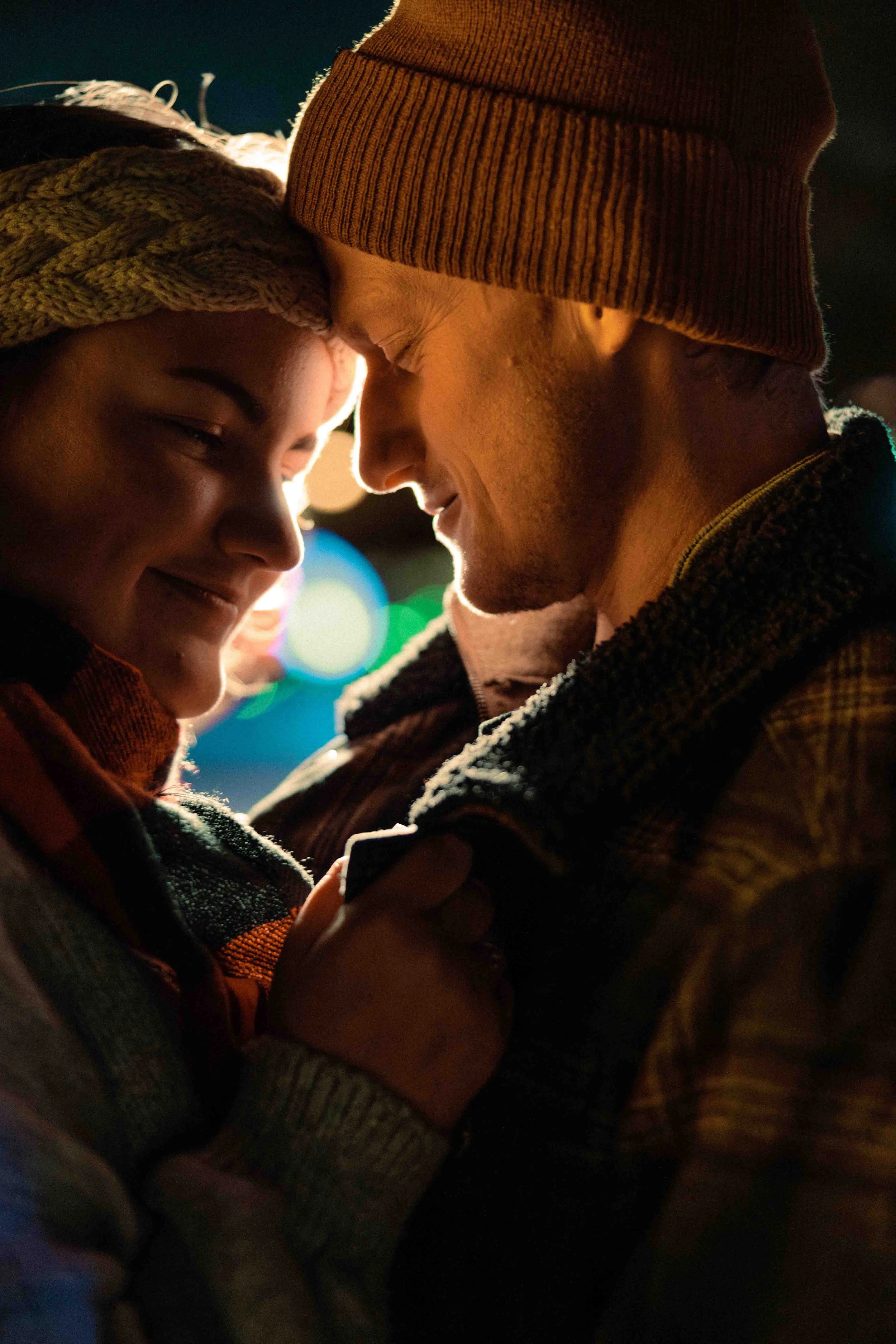 A couple smiling and leaning their foreheads together at night, with colorful lights blurred in the background.