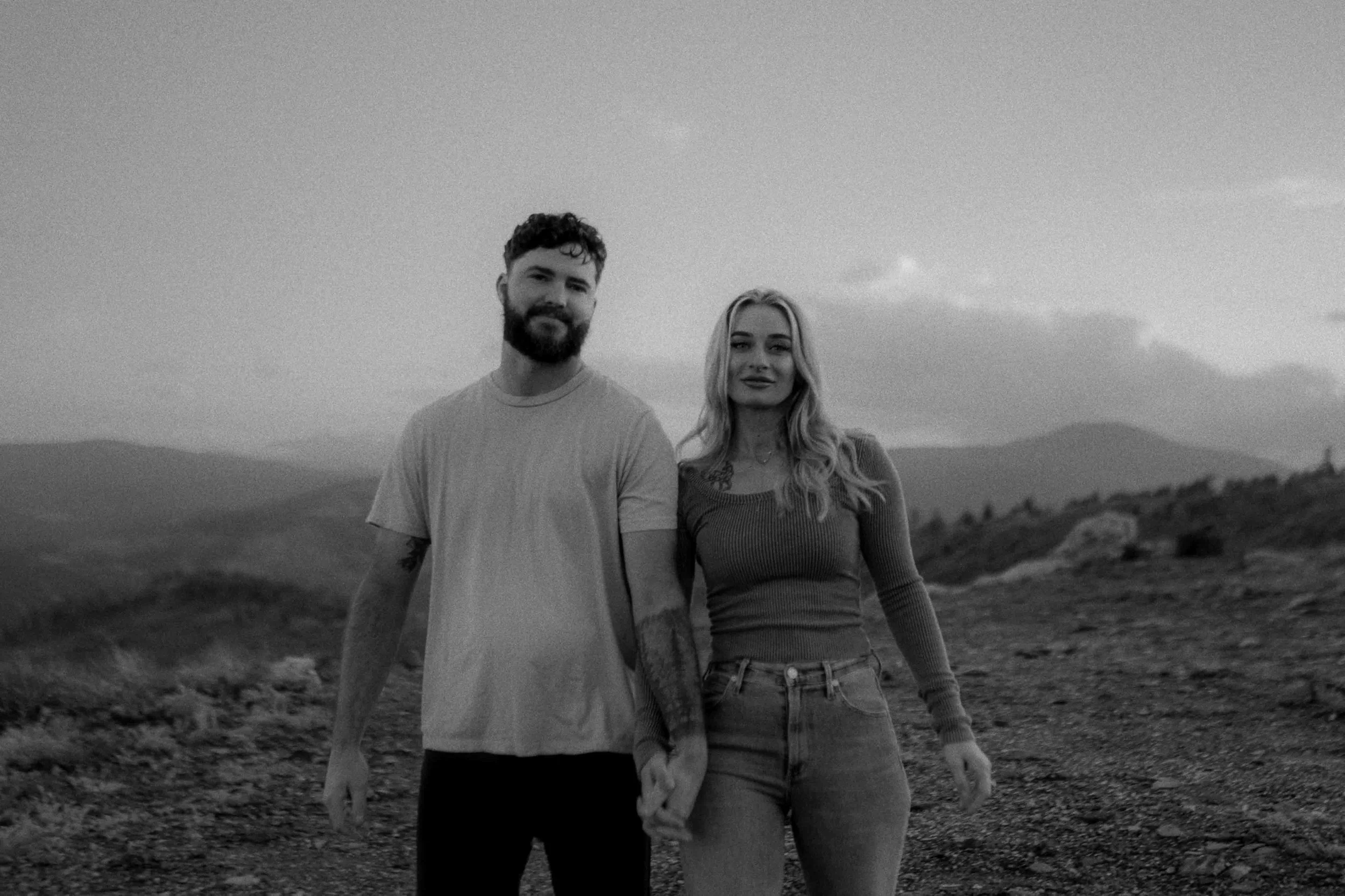 A black and white photo of a young man and woman holding hands outdoors with mountains in the background.