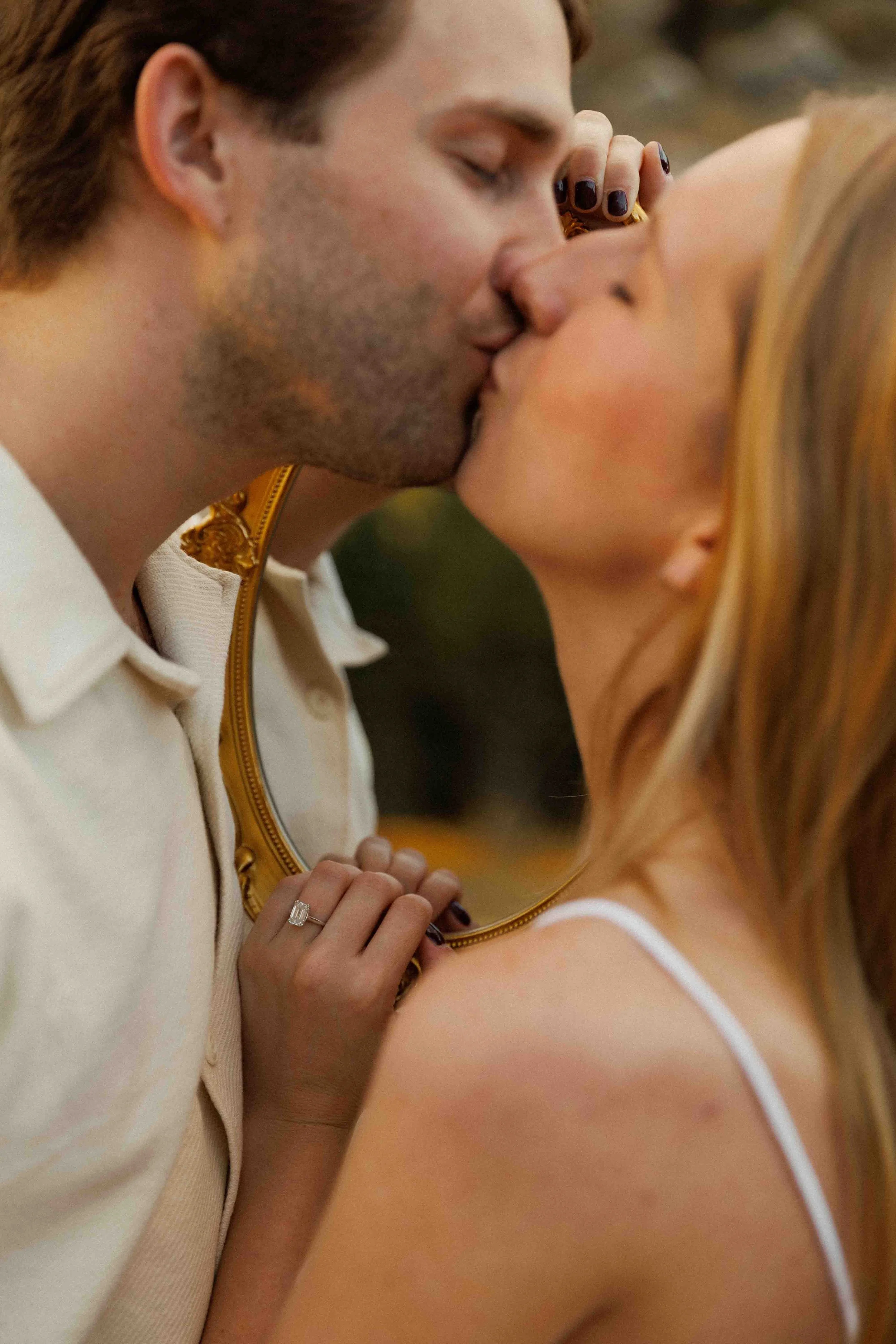 A couple sharing a kiss, with the woman holding a mirror reflecting her face, and her hand displaying an engagement ring.