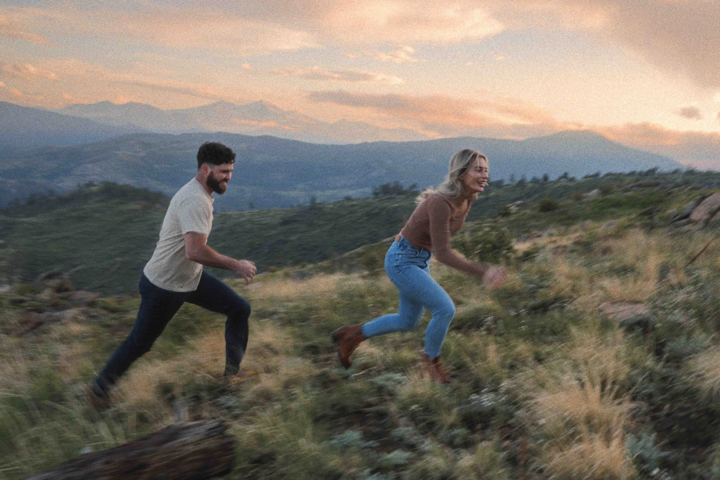 A man and woman running through a grassy field at sunset with mountains in the background.
