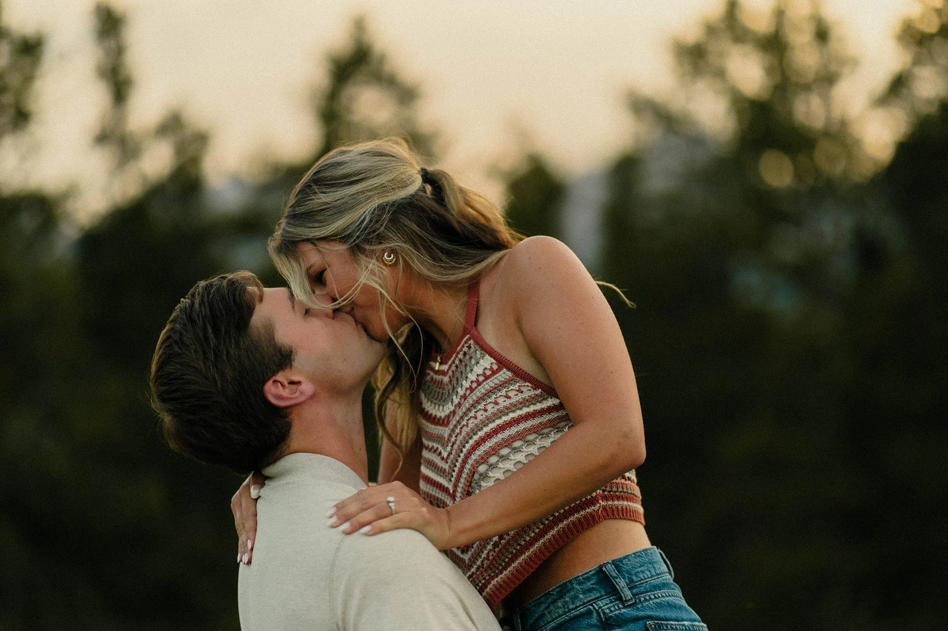 A couple kissing outdoors during sunset with blurred trees in the background.