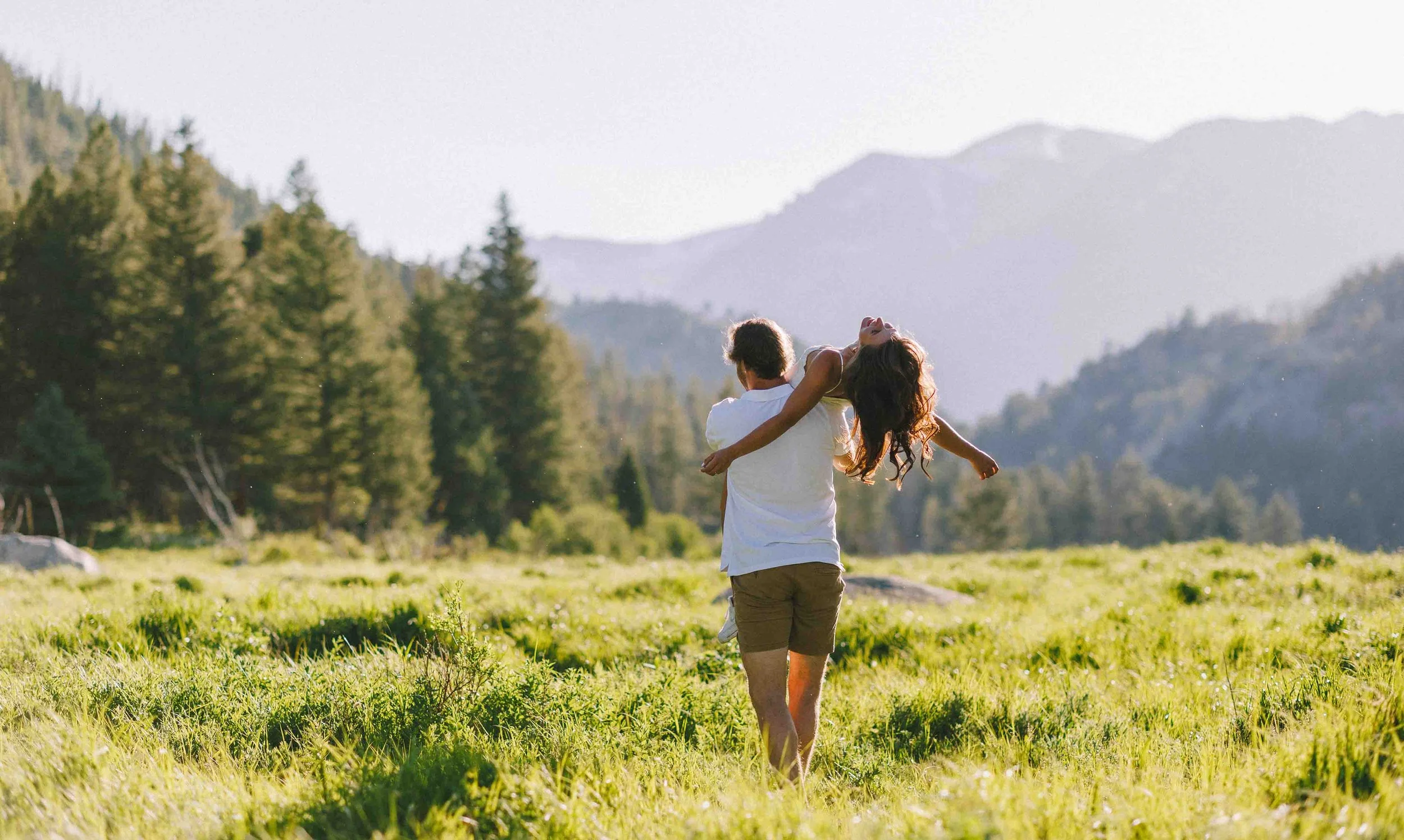 A person holding a woman in their arms in a grassy field with trees and mountains in the background during daylight.