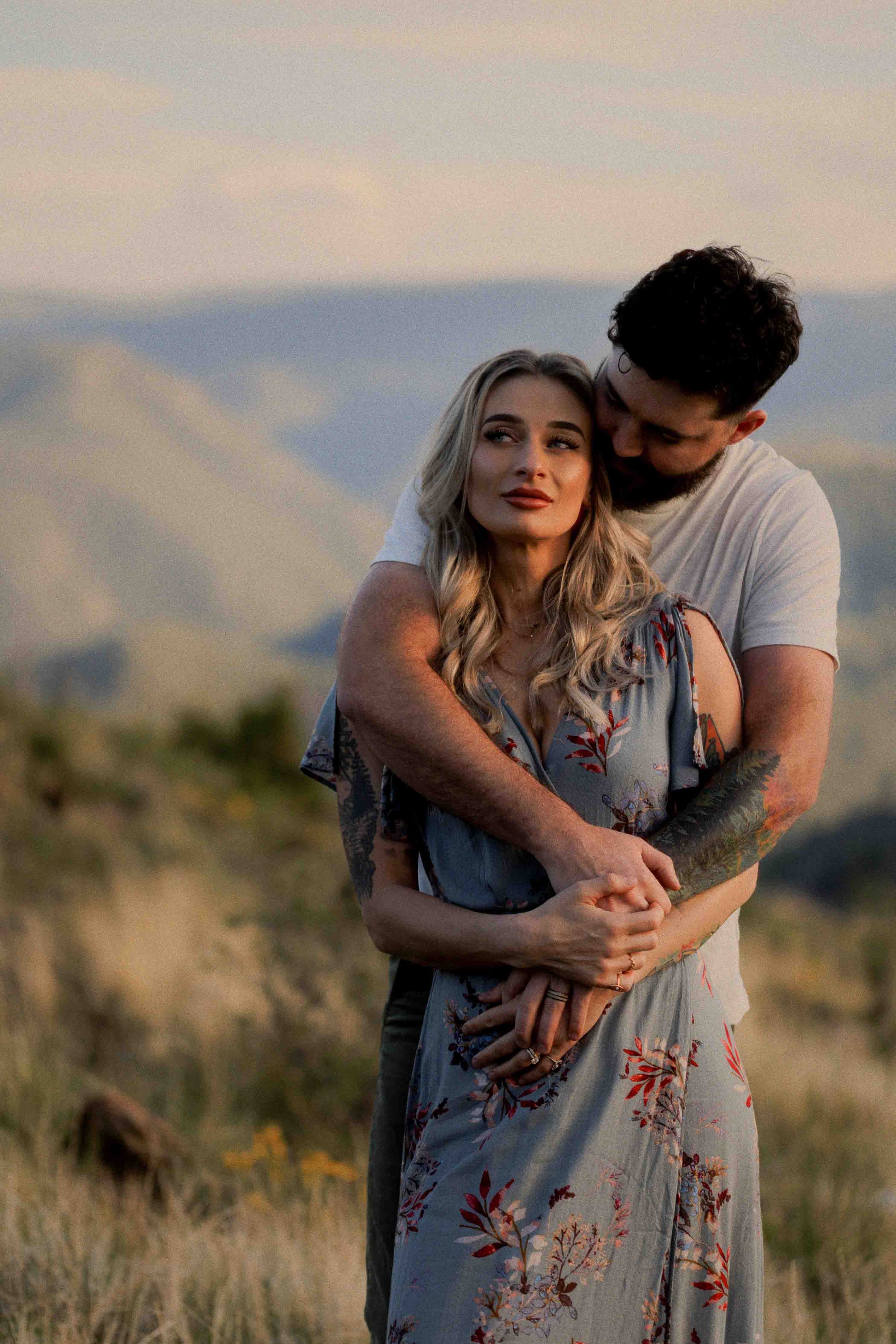 A couple embracing outdoors during sunset, with mountains and a rainbow in the background.