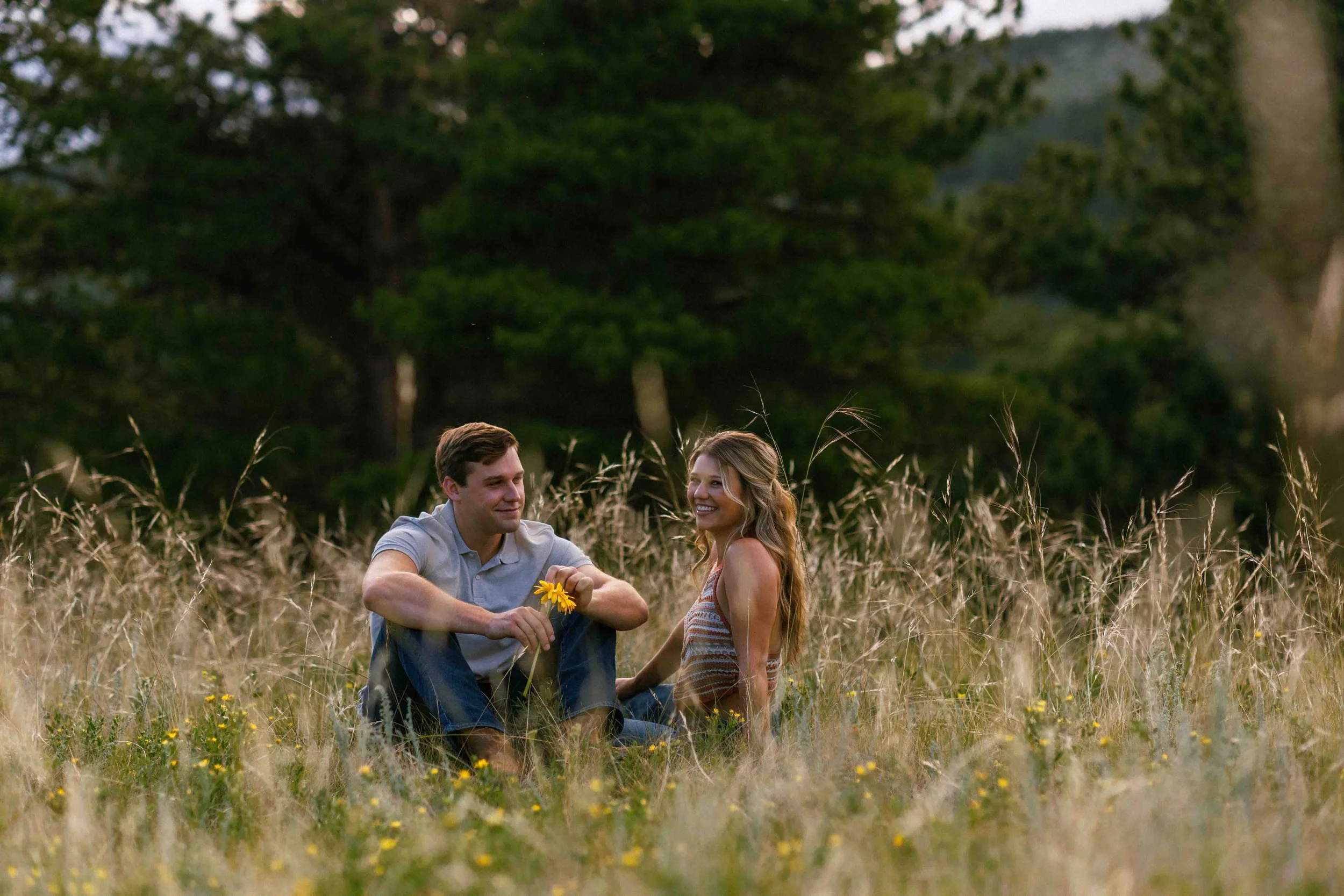 A young man and woman sitting in a grassy field with yellow flowers, trees in the background, during sunset, sharing a moment together.