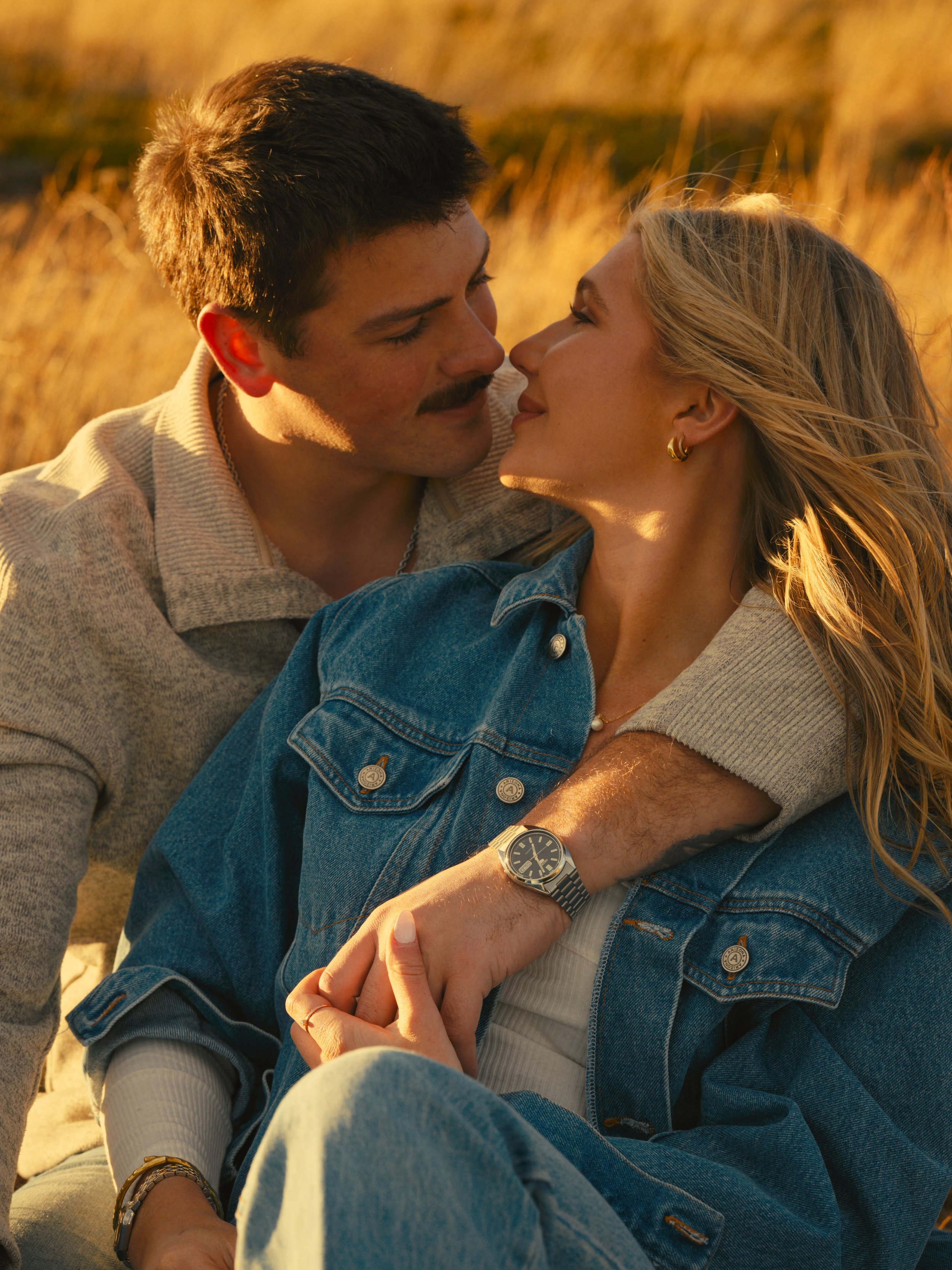 A man and woman sitting closely together outdoors during sunset, with the man holding the woman's hand and sharing an intimate moment.
