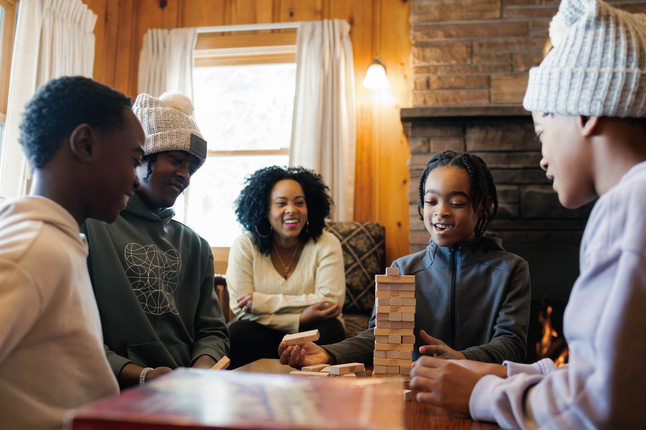 Group of people playing a block-stacking game inside a cozy room with wood walls and a fireplace. One person is holding a block, while others watch and smile.