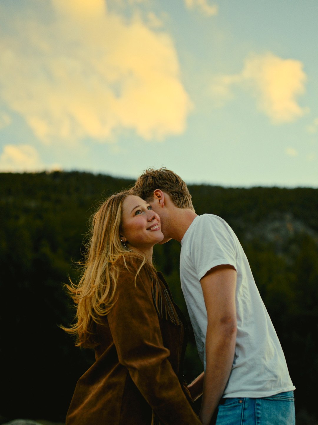 A couple standing outdoors, the woman looking up with a smile, while the man kisses her on the cheek, with a mountain and cloudy sky in the background.