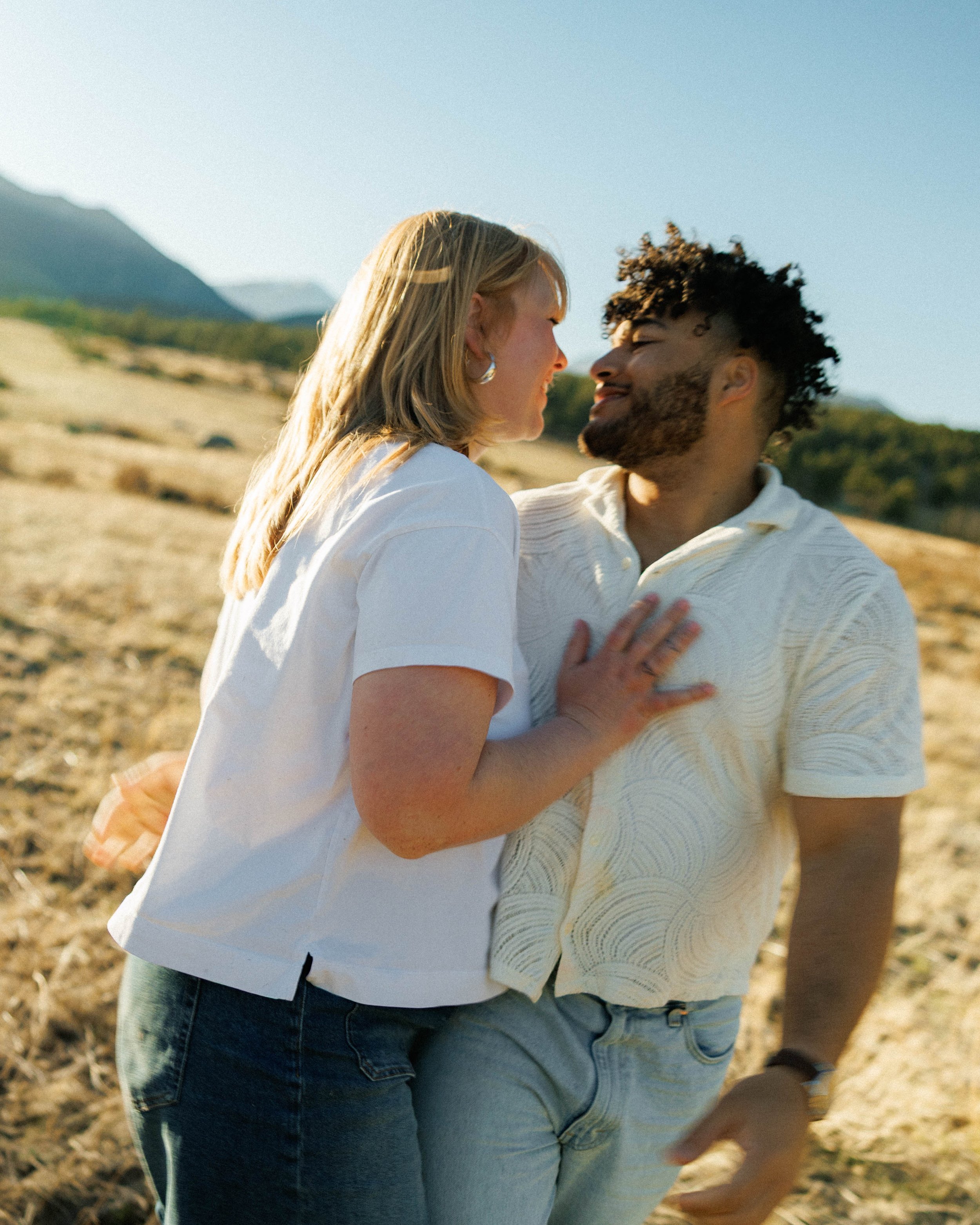 A couple smiling and embracing outdoors in a sunny, mountainous landscape.