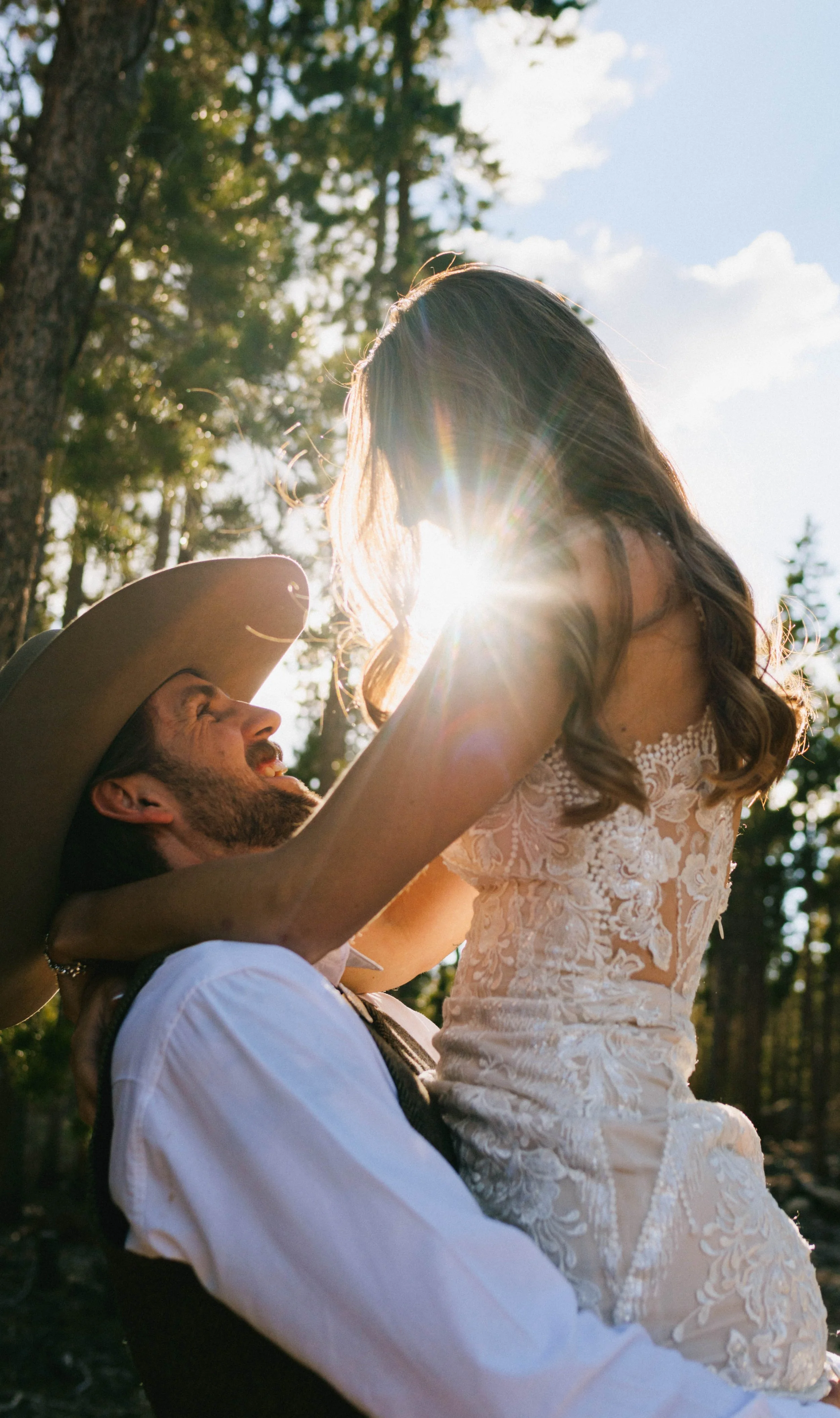 Happy couple embracing outdoors, man wearing cowboy hat, woman in lace dress, sunlight filtering through trees. Elopement engagement wedding shoot