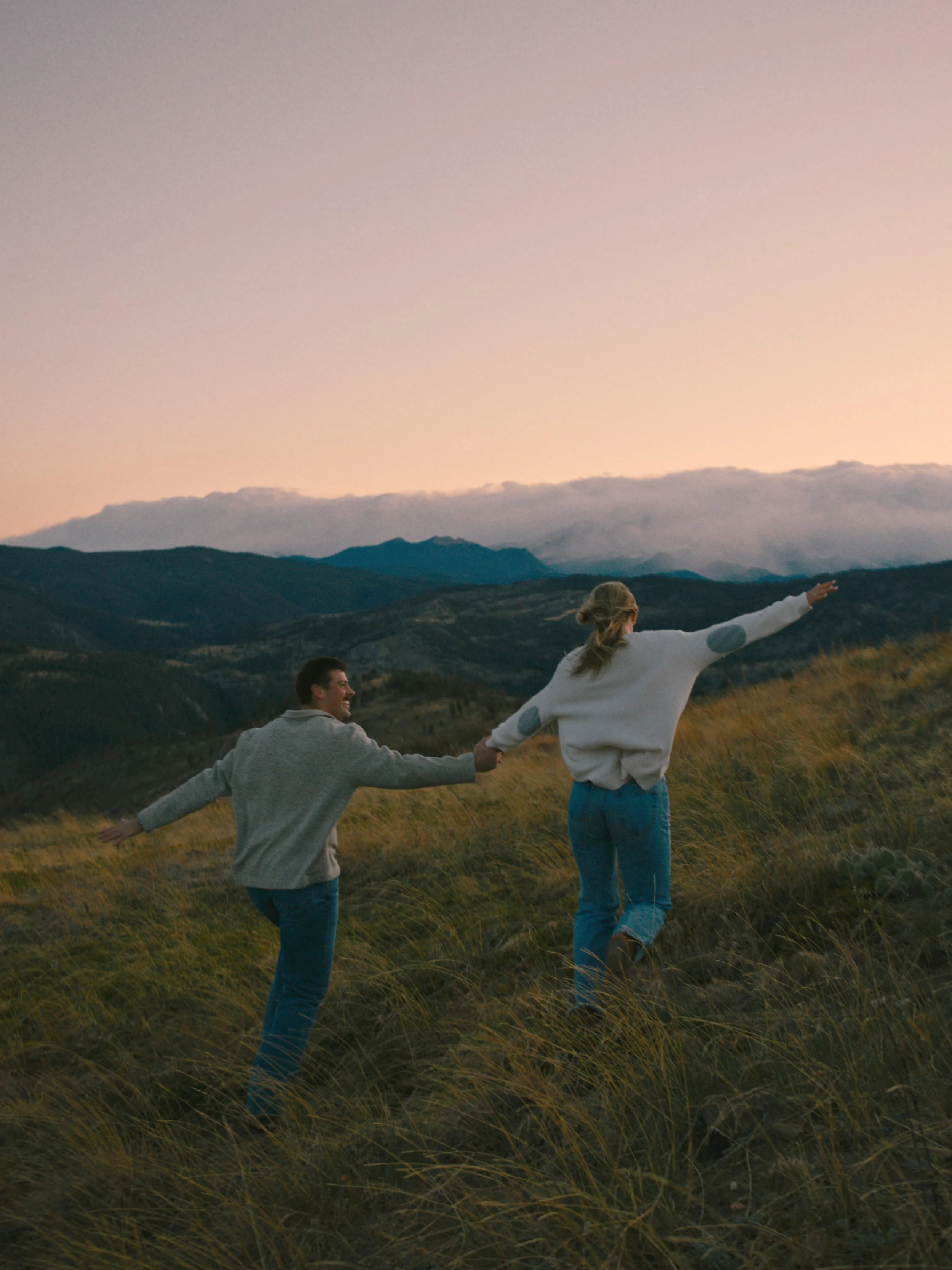 A couple holding hands and playing in a grassy field at sunset with mountains in the background.