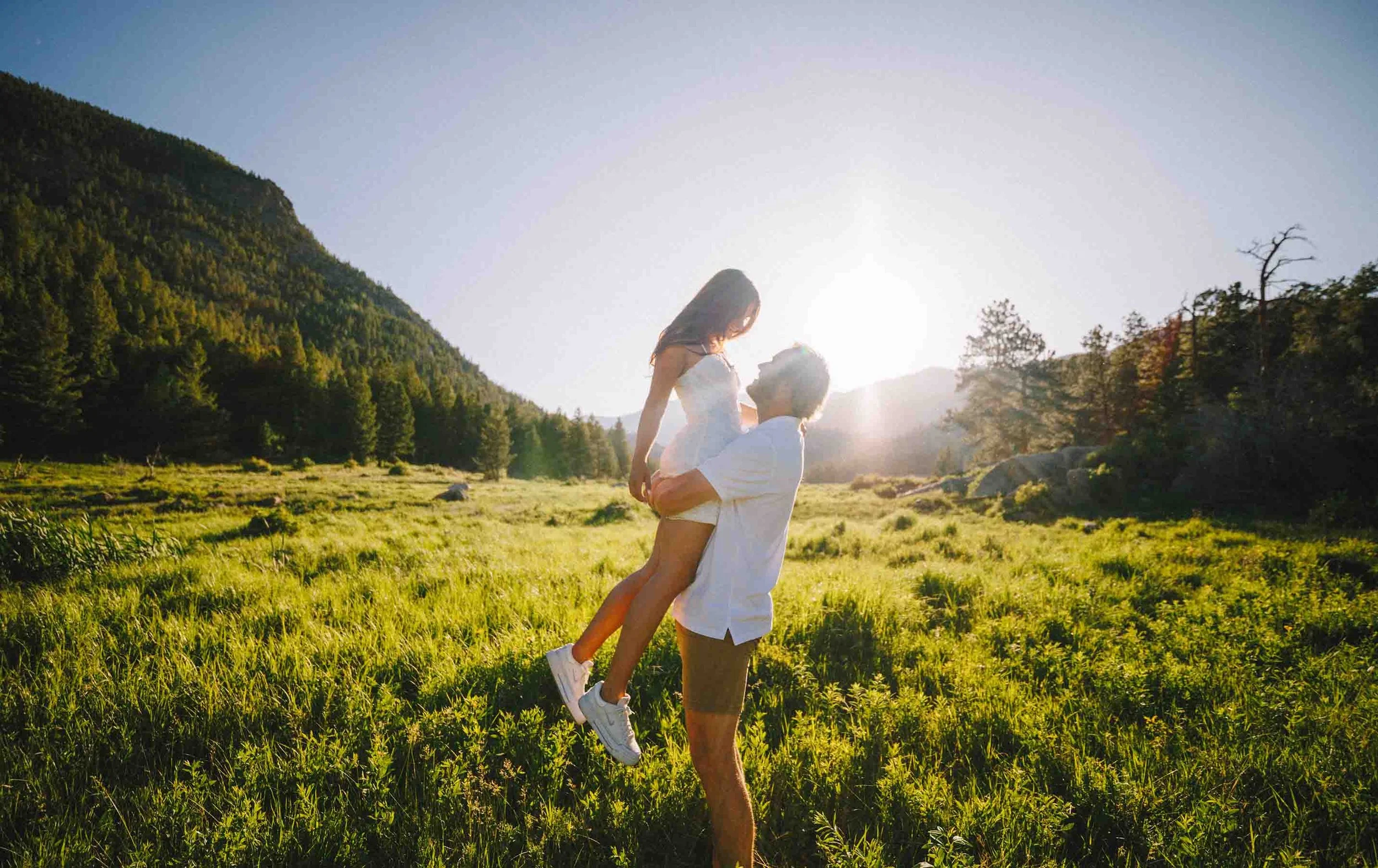 A man lifting a woman in a white dress in a lush green field during sunset, with mountains and trees in the background.