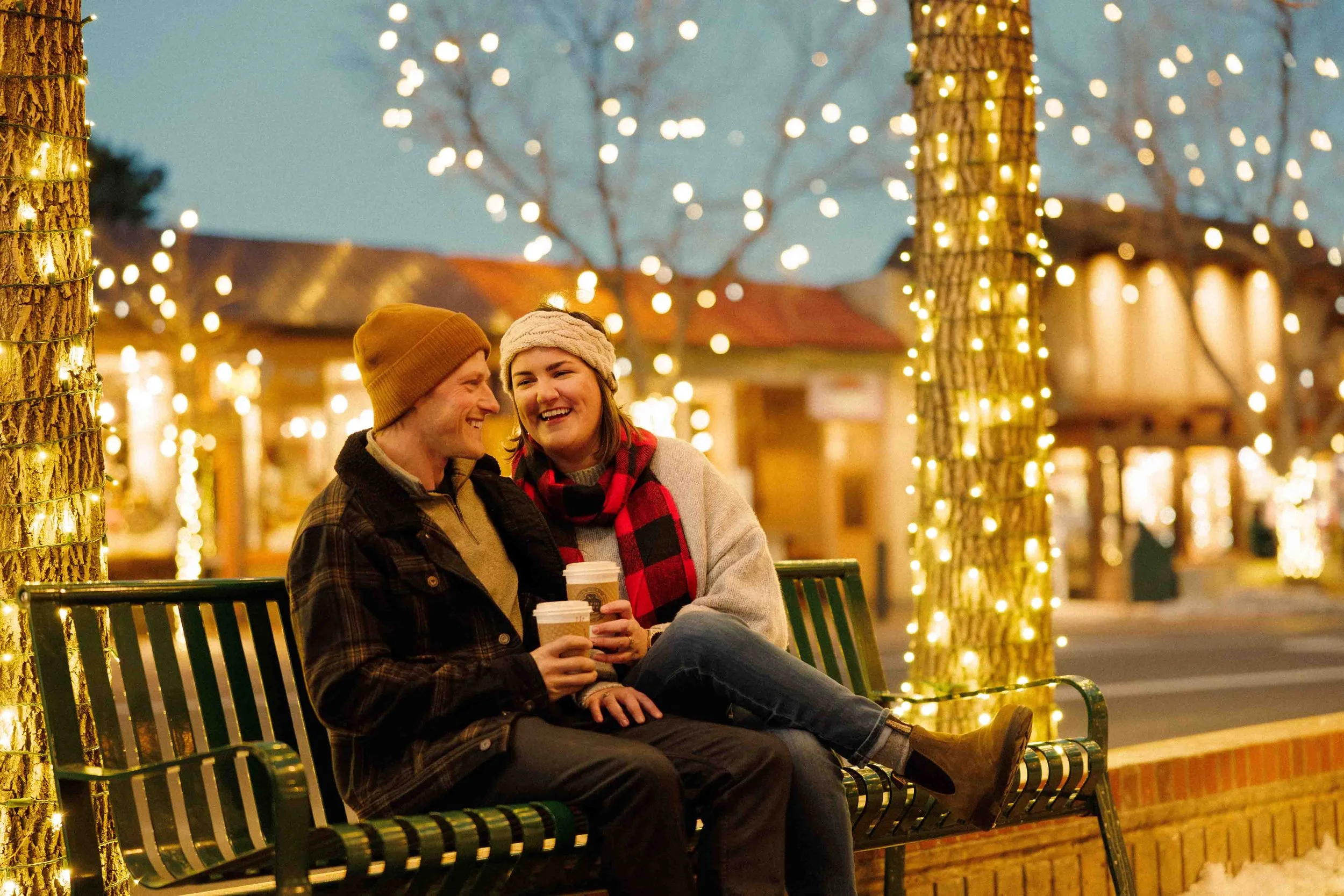 A couple sitting on a bench at night, holding coffee cups, surrounded by Christmas lights wrapped around trees, with warm orange and yellow lights illuminating a street scene.