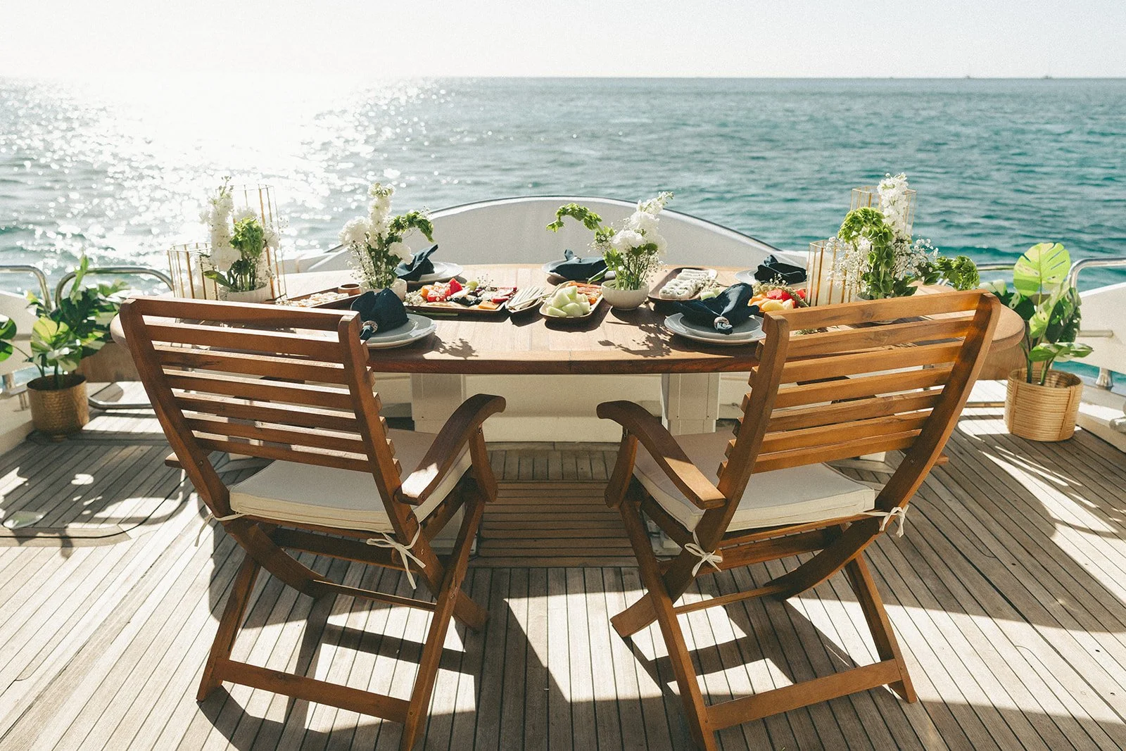 A dining table set for a meal on a boat, with four wooden chairs and a centerpiece of white flowers, overlooking the ocean.