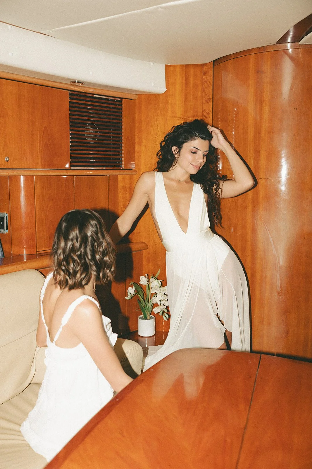 A woman in a white dress standing and smiling, while a girl in a white dress with curl hair sits on a beige sofa with a flower vase on a wooden table nearby in a room with wood-paneled walls.
