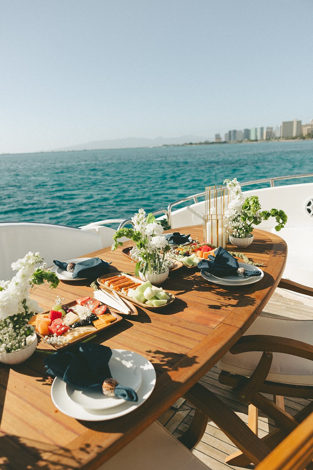 A wooden dining table set on a yacht deck with plates of food, flowers, and decorations, overlooking a body of water and distant city skyline.