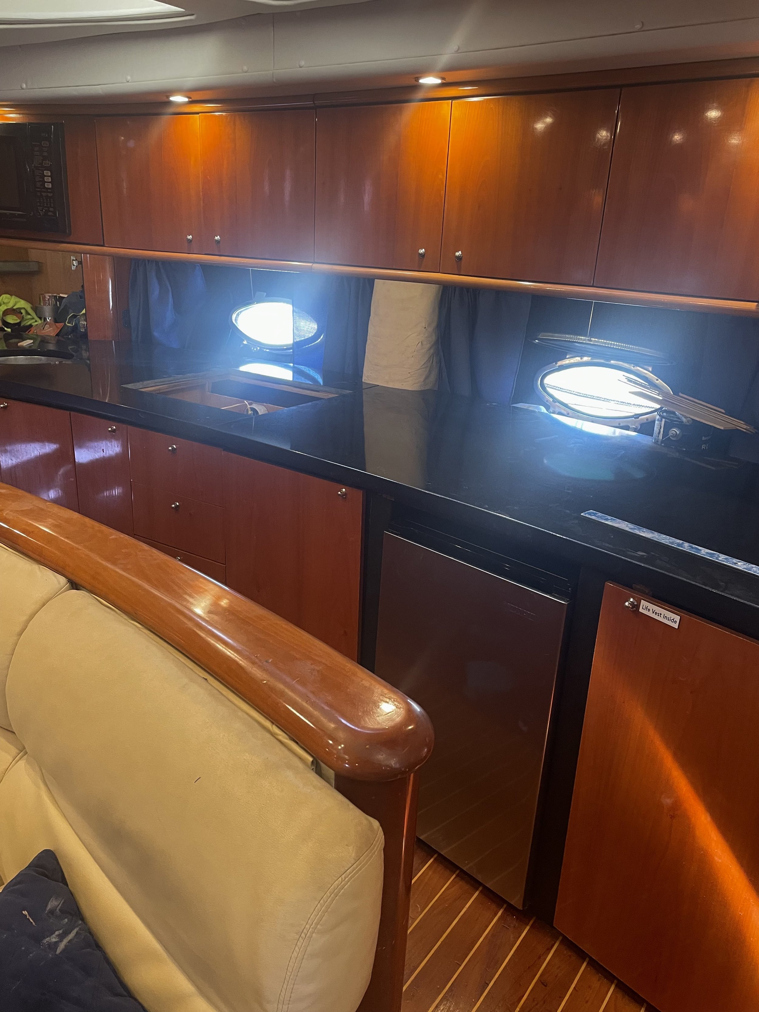 Interior of a boat or yacht with wooden cabinets and a black countertop. There are small circular portholes with sunlight coming through. A beige cushioned seating area is visible at the bottom of the image.