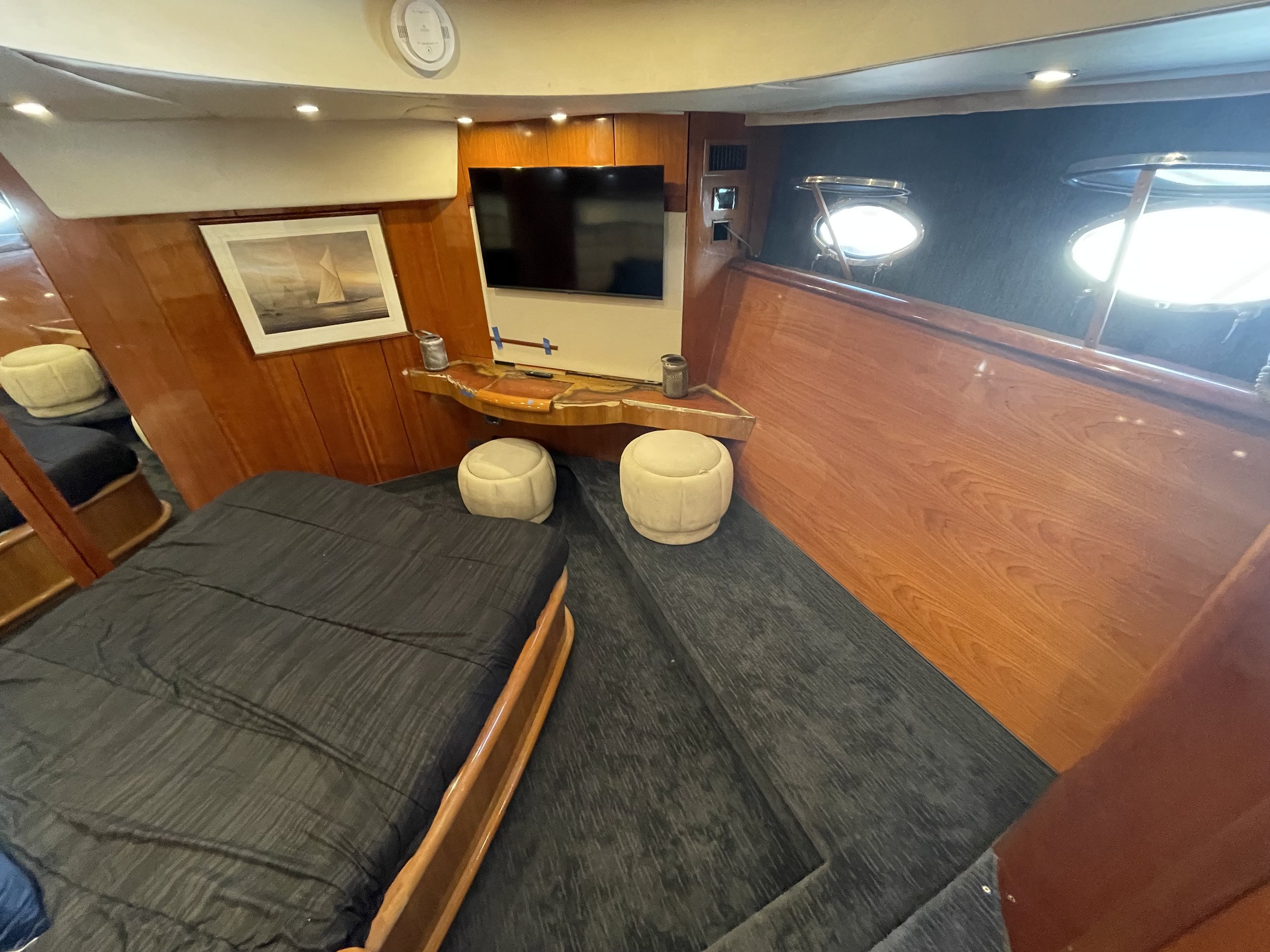 A room with a flat-screen TV mounted on a wood-paneled wall, a chair, a framed ocean-themed artwork, and a porthole window with sunlight coming through.