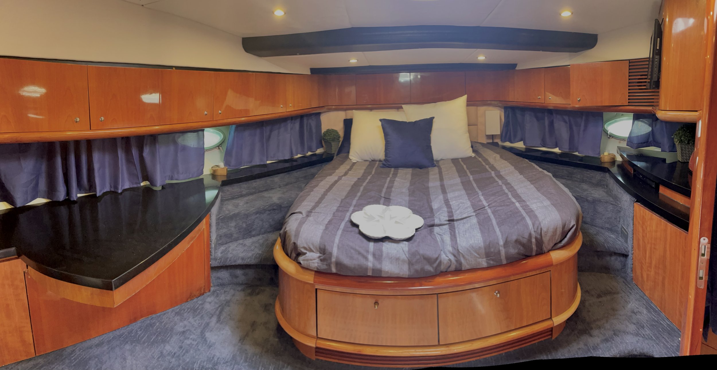 A cozy bedroom with a round bed covered in a gray striped bedspread, decorative pillows, dark purple curtains beneath small porthole windows, wooden cabinetry above, and a black countertop to the side with small decorative items and plants.