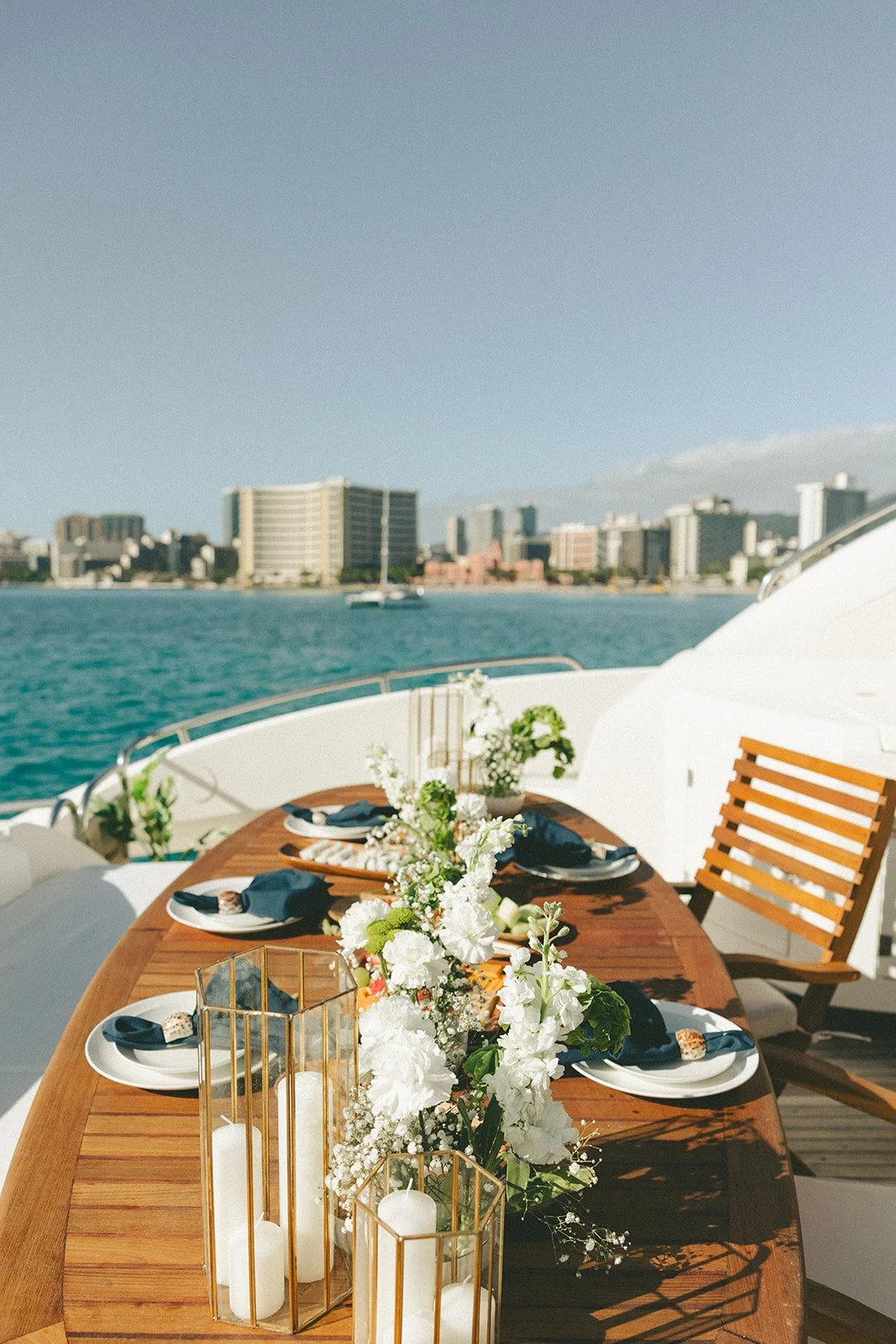 Set dining table on a yacht with a city skyline in the background, decorated with white flowers and candles, with blue napkins on plates.