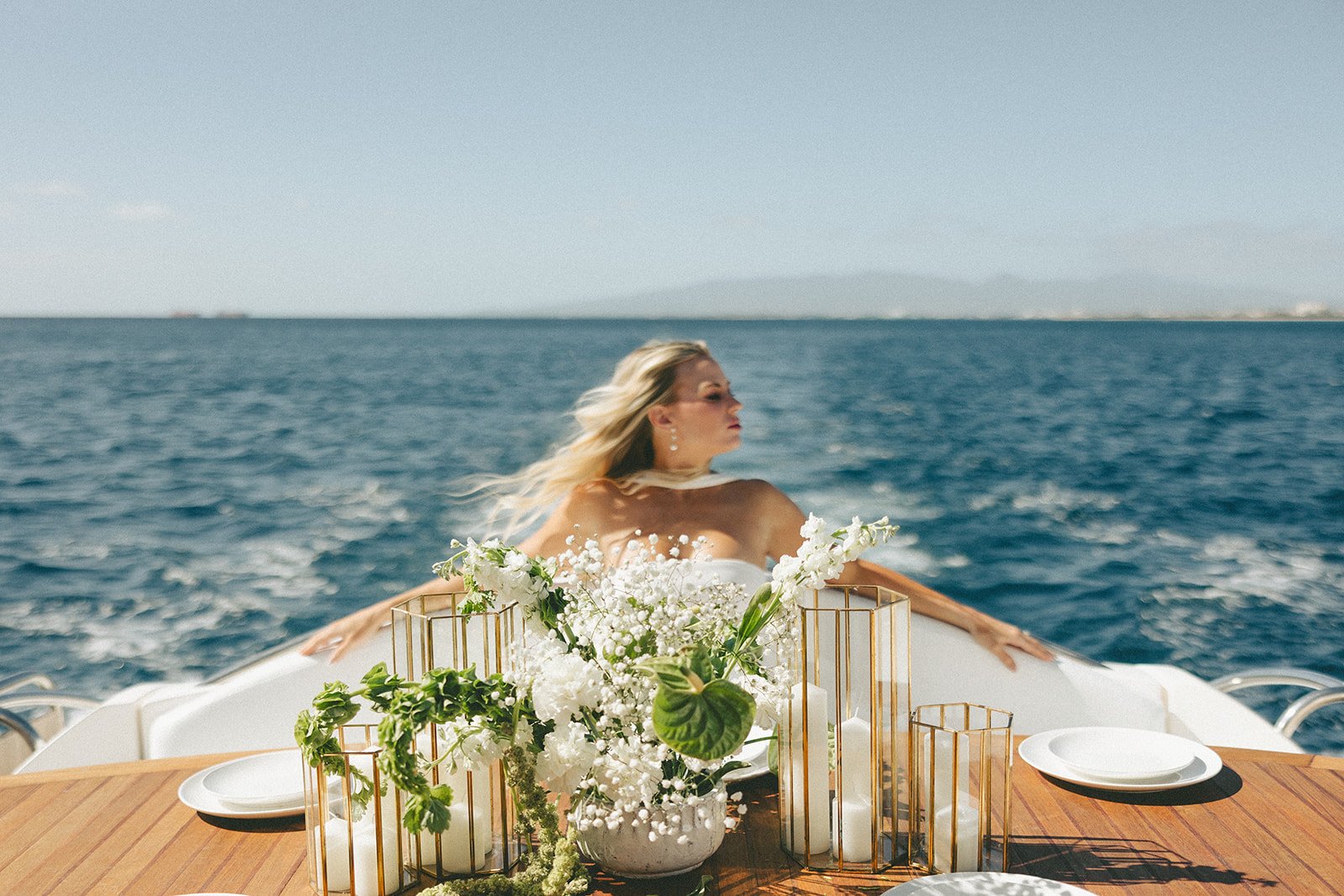A woman with blonde hair and earrings is on a boat, leaning on the back, with the ocean and a distant shoreline in the background. The boat's table in front of her is decorated with a white floral arrangement and gold candlesticks.