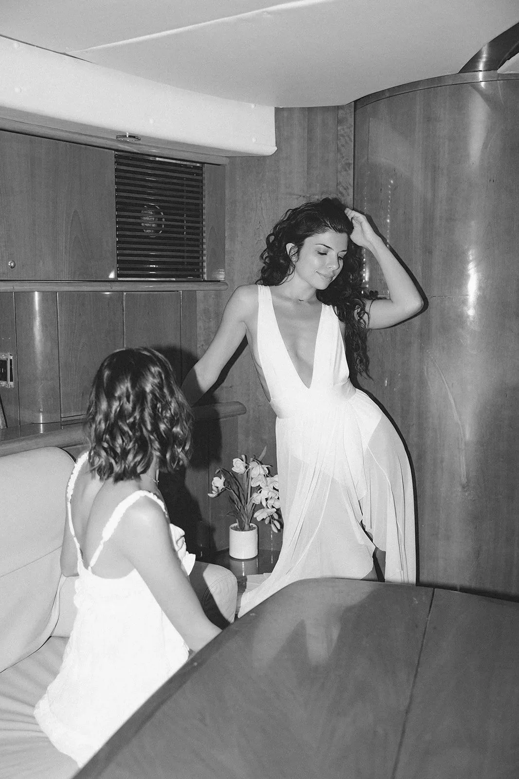 A woman in a white dress leaning against a wall with her eyes closed, while a young girl with curly hair in a white dress sits on a bench nearby, in a room with wooden paneling and a potted plant.