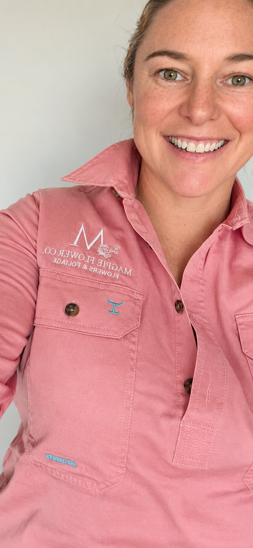 A woman smiling, wearing a pink collared shirt with embroidery that reads "Magpie Flower Co., Flowers & Foliage" and a small white logo.