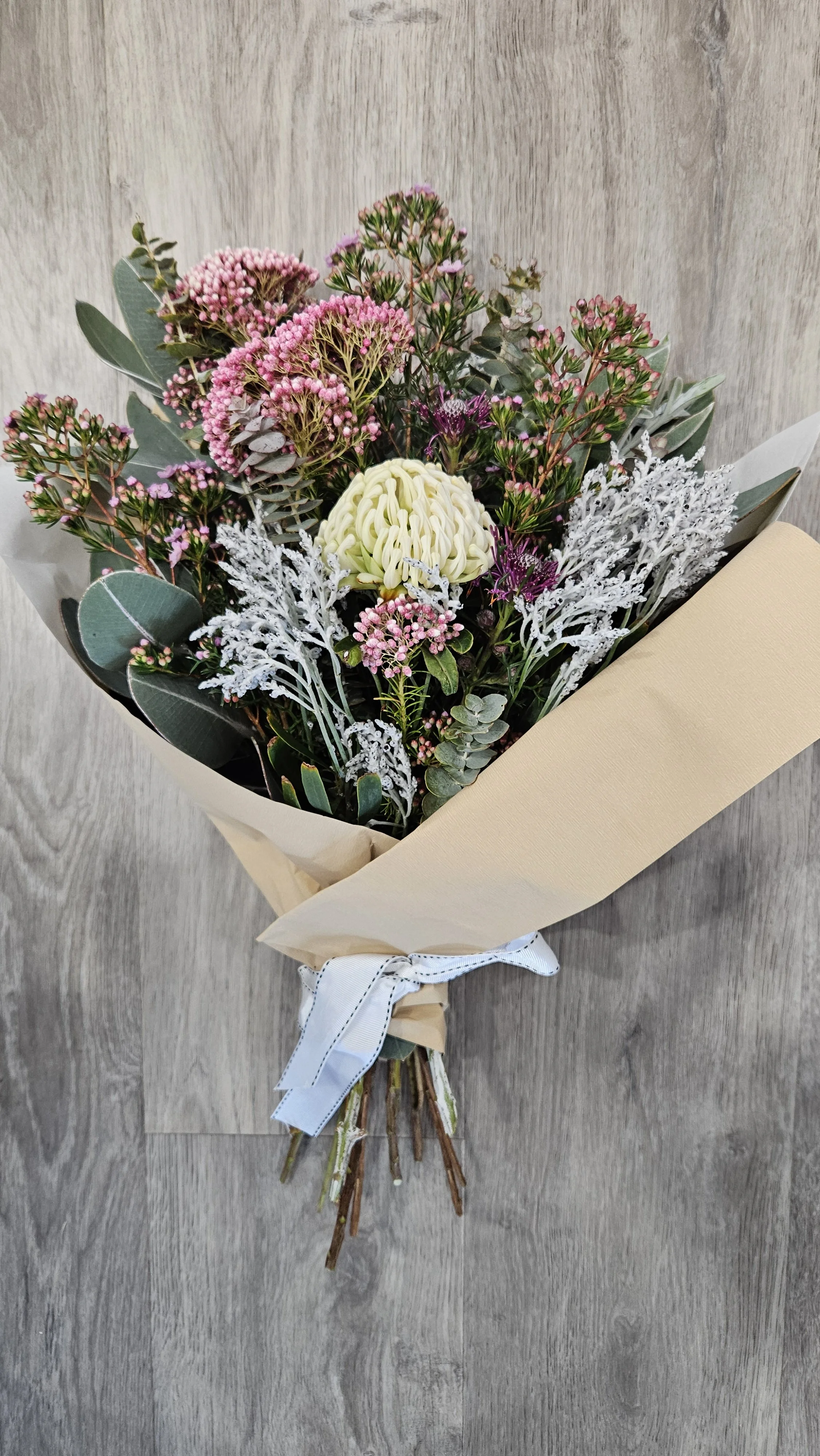 A bouquet of pink, purple, and white flowers with green foliage, wrapped in beige paper and tied with a white ribbon, resting on a wooden surface.